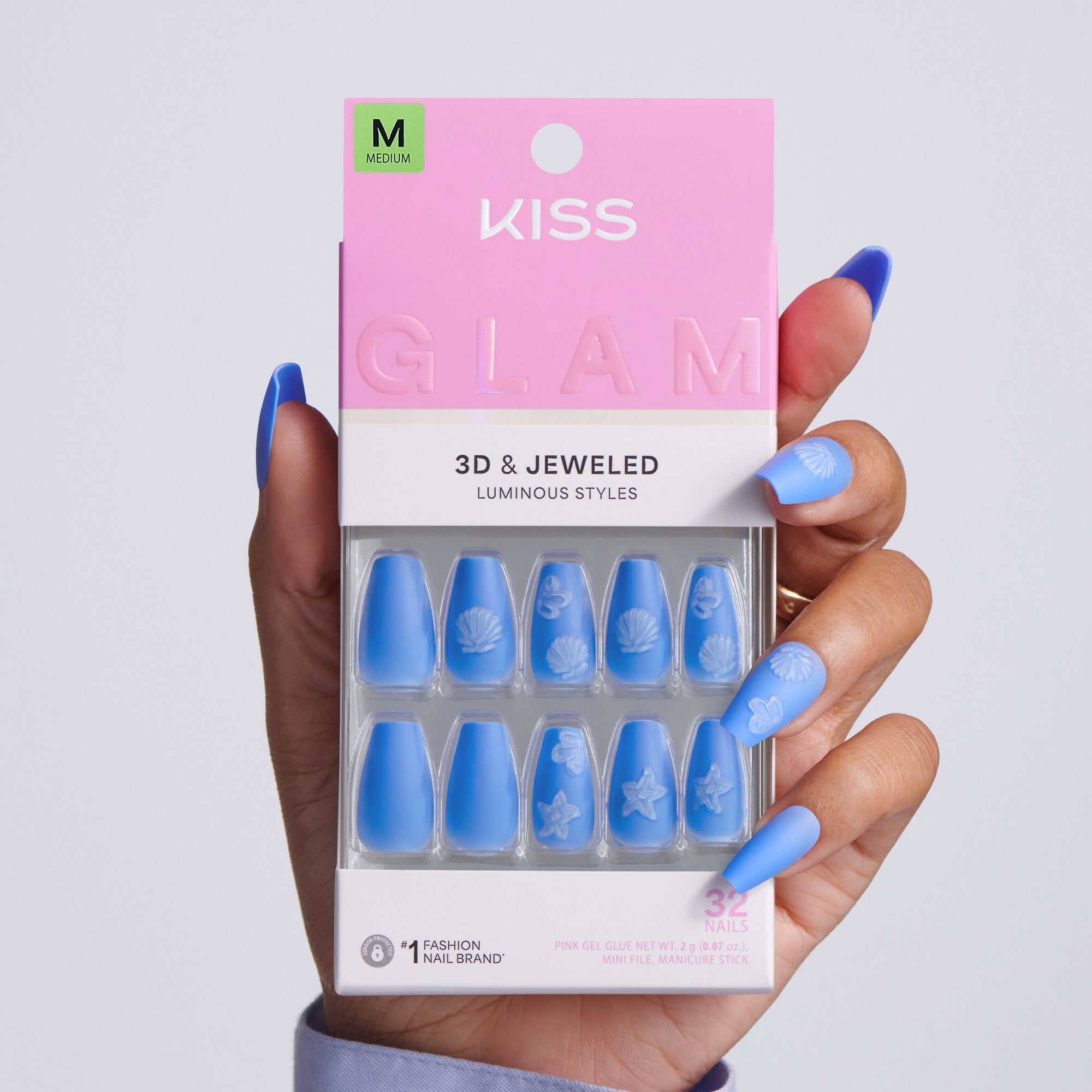 Hand holding KISS Glam 3D & Jeweled Luminous Styles press-on nails packaging, Medium size, 32 nails, blue medium coffin nails with white embossed shell and ocean designs on gray background