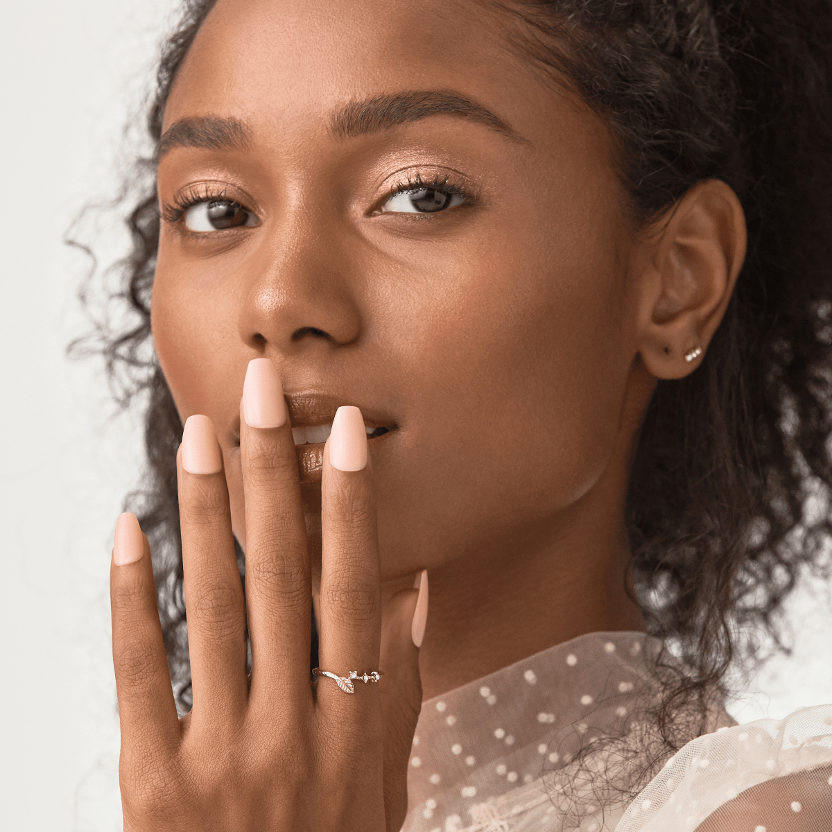 Woman with medium brown skin wearing matte pale peach press-on nails from imPRESS Color Press On Nails in the Serendipity shade, with a delicate ring and sheer polka dot top.