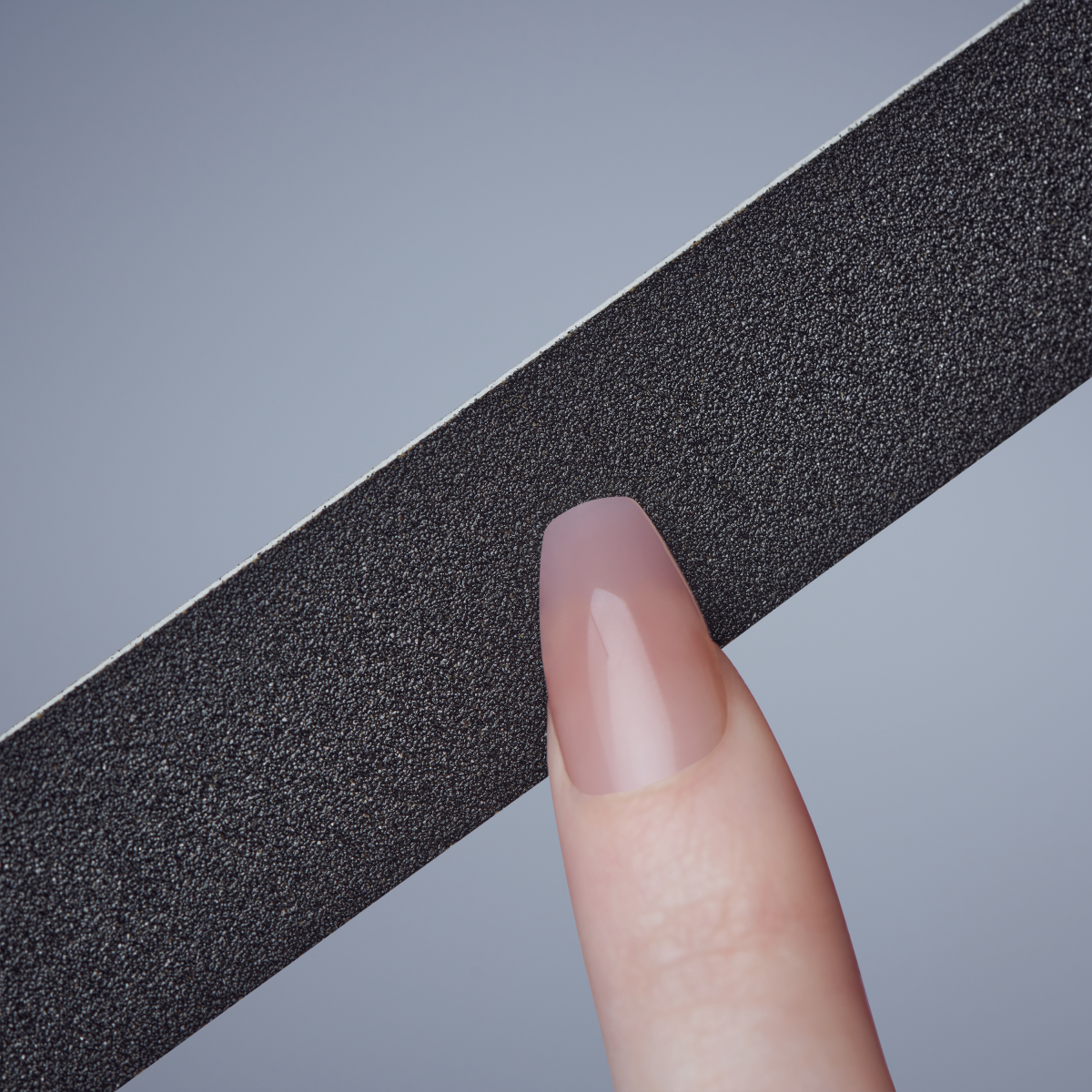 Black textured nail file held horizontally against a grey background, with a close-up of a manicured finger showcasing a smooth, glossy, nude-colored nail.