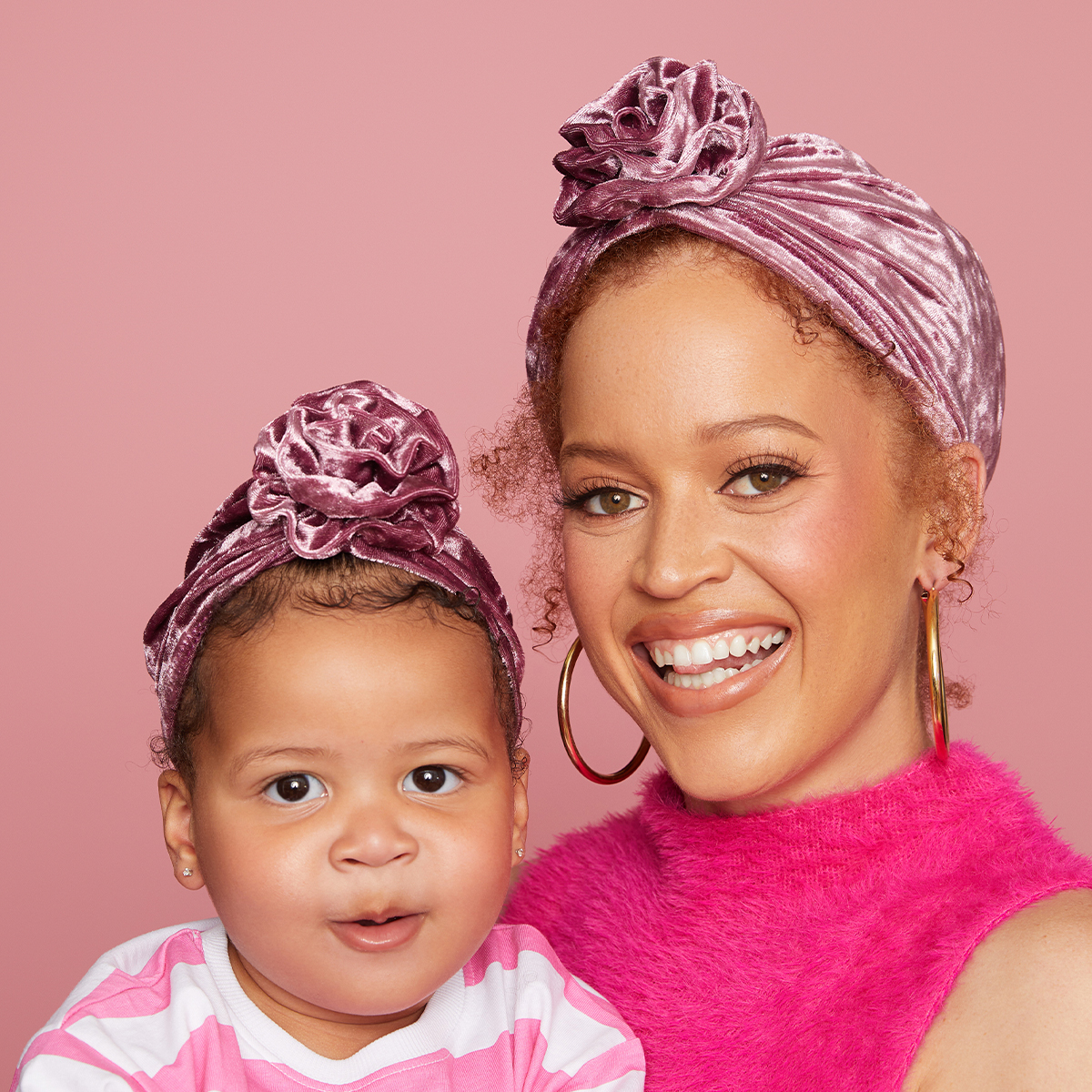 Smiling woman and toddler wearing matching soft berry-colored velvet pre-tied top knot turbans against a pink background.