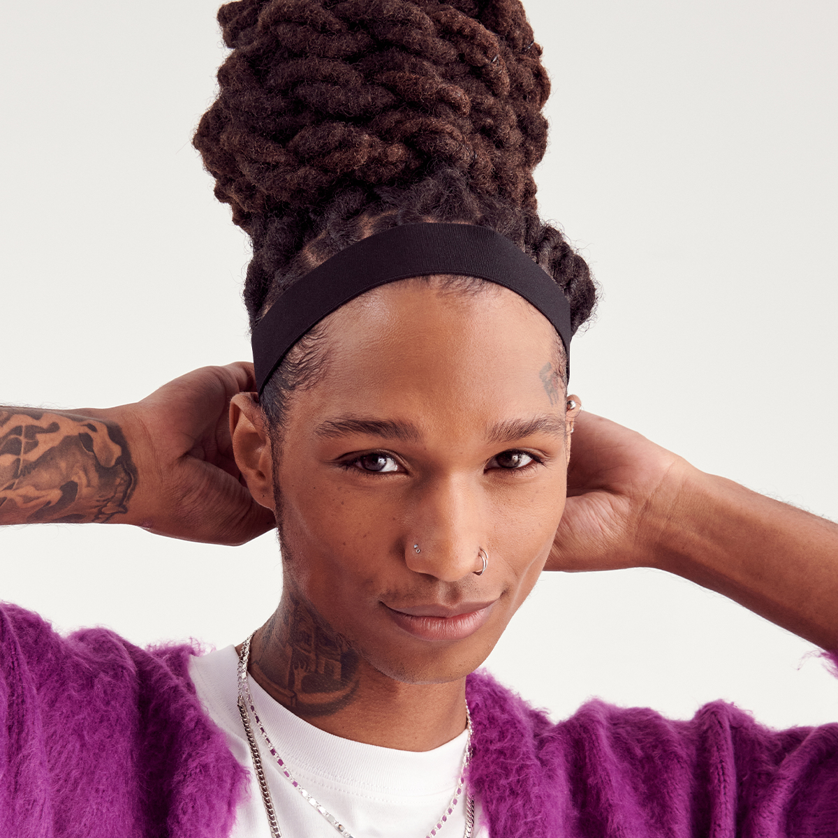 Person with long braided hair styled in a high bun wearing a black elastic headband, purple fuzzy cardigan, and silver necklaces against a plain background.
