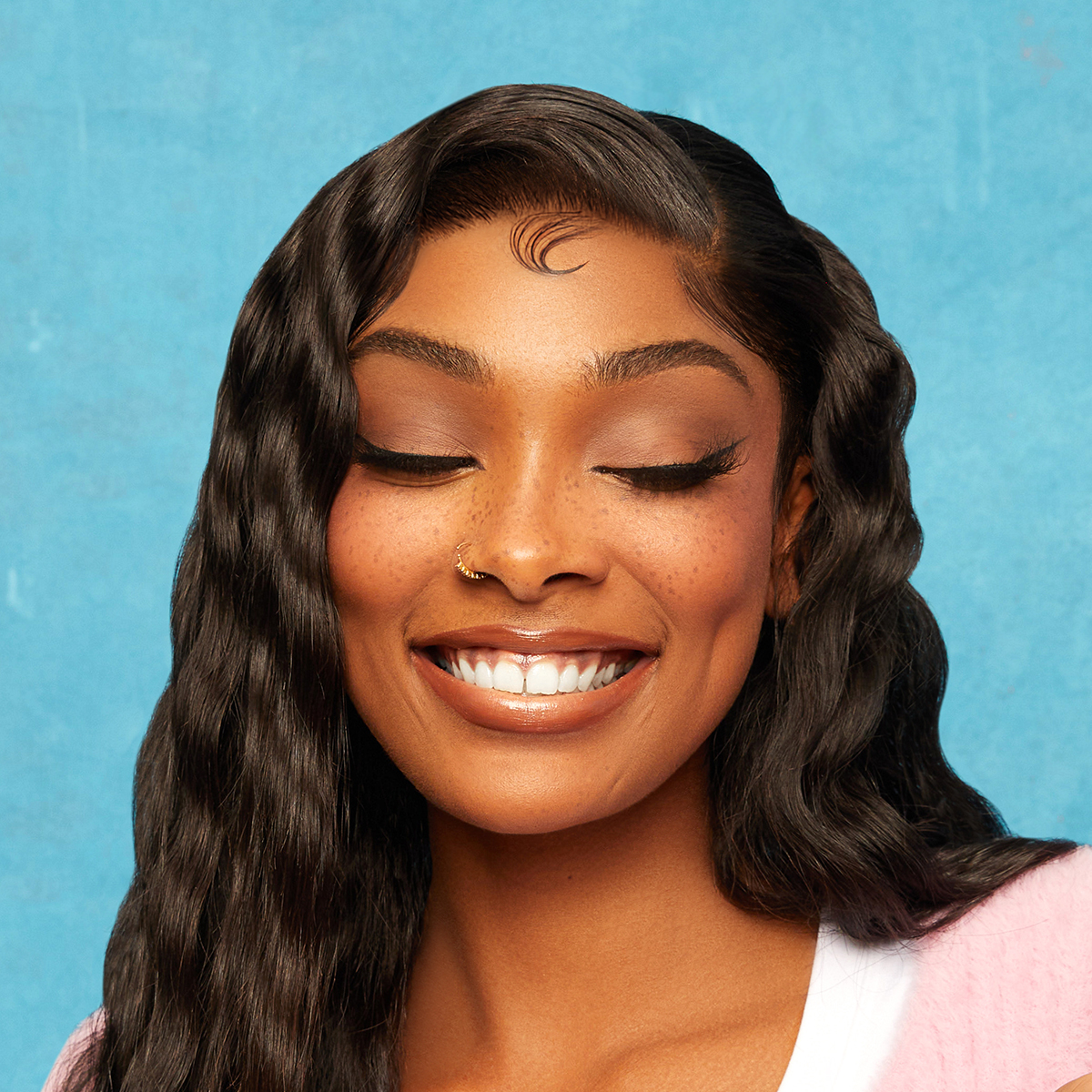 Woman with smooth, styled black wavy hair and neatly laid edges, smiling against a blue background, wearing a pink sweater over a white shirt.