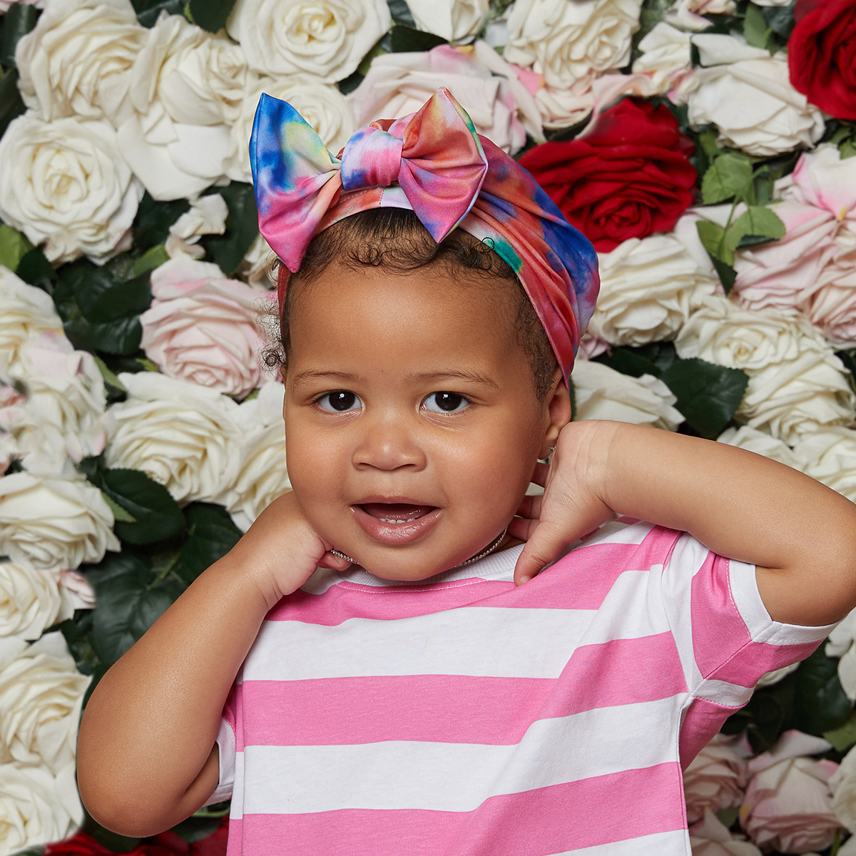 Toddler wearing a colorful tie-dye pre-tied top knot bow turban wrap, standing in front of a backdrop of mixed white and red roses.