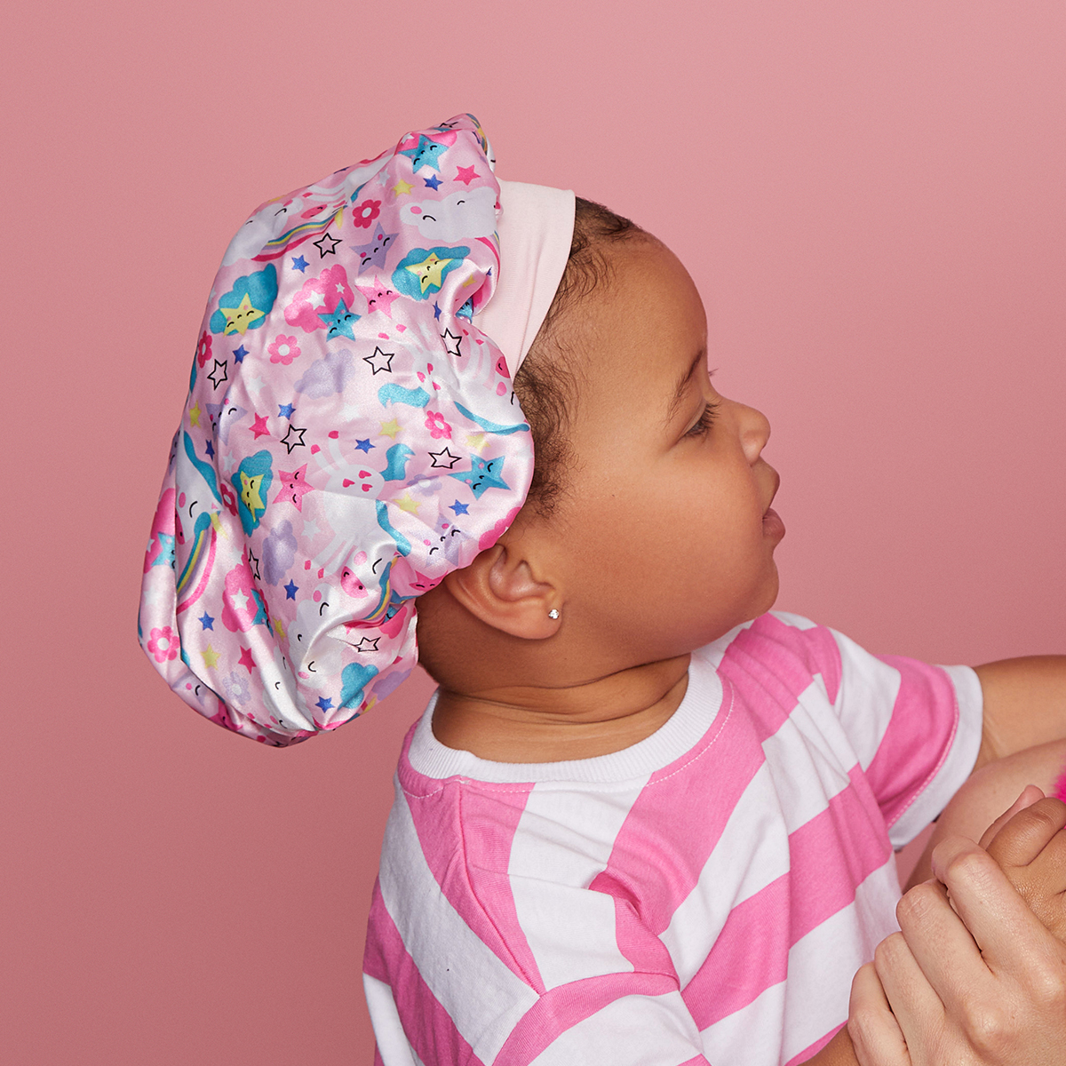 Toddler wearing a silky satin bonnet with a colorful pattern of stars, clouds, flowers, and rainbows, against a pink background.