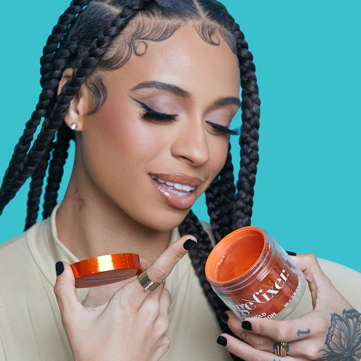 Young woman with braided hair and winged eyeliner holds an open 100 mL jar of orange KISS Colors & Care Edge Fixer, smiling against a turquoise background.