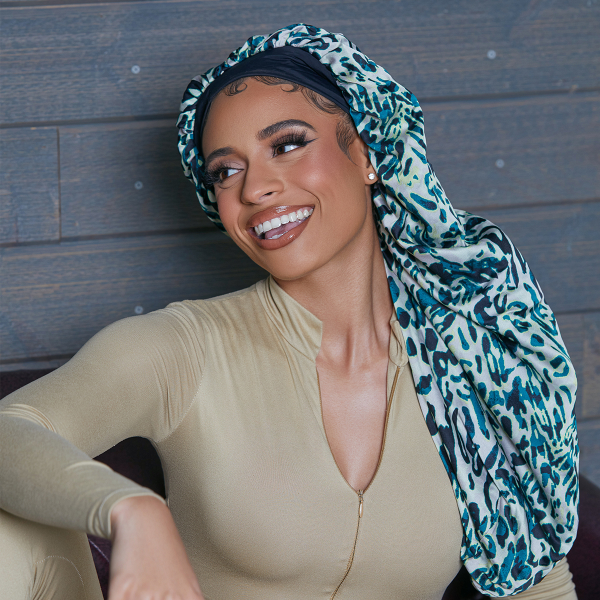 Woman smiling and looking to the side wearing a crepe satin reversible braid bonnet in an exotic blue and black leopard print, paired with a beige zip-up outfit.