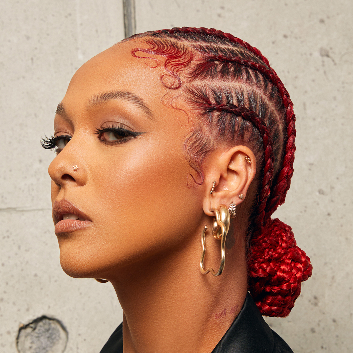 Woman with bright red braided hair styled in cornrows and edges neatly styled in sleek waves, wearing multiple gold earrings and a black top against a concrete background.