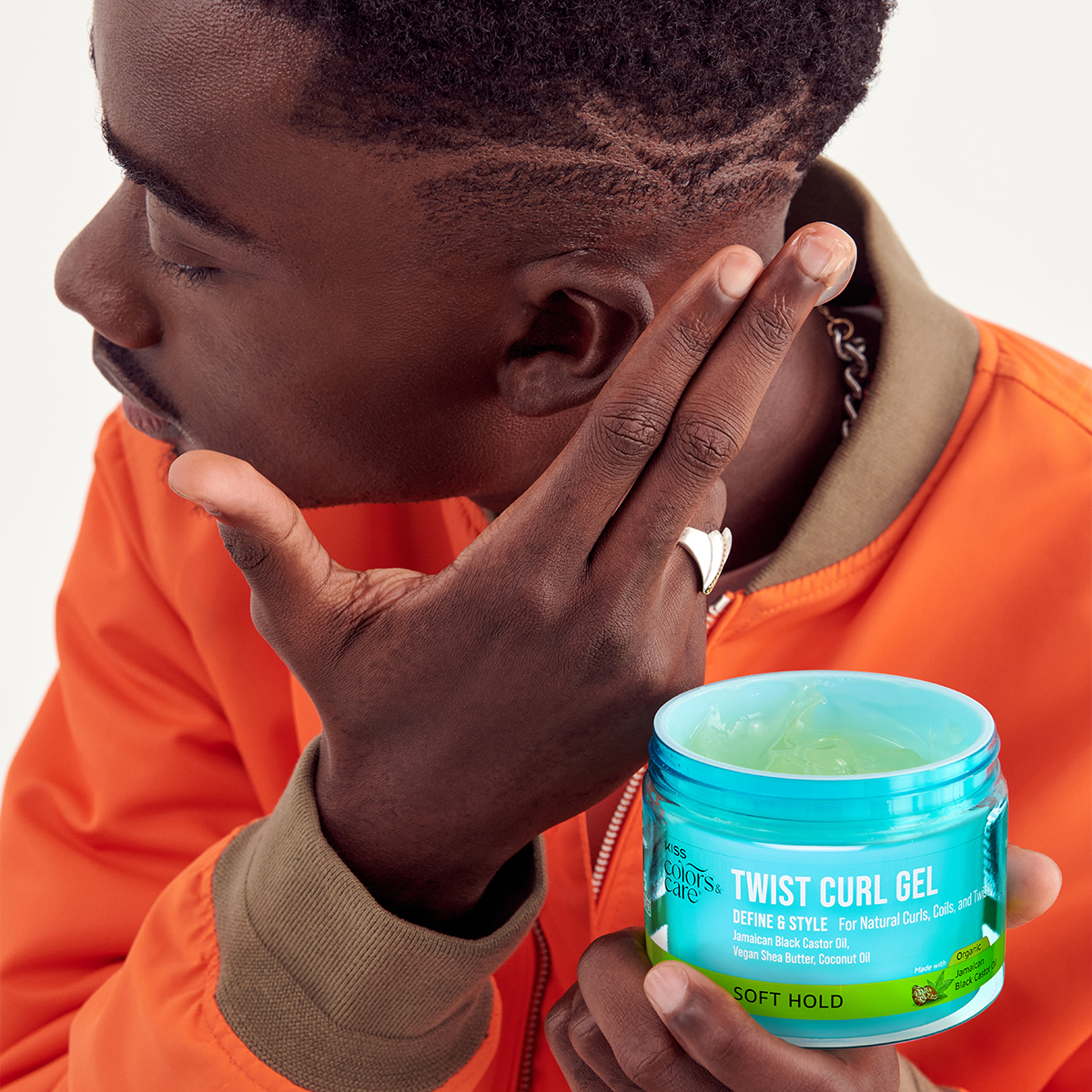 Man wearing orange jacket applying clear gel from a blue container labeled KISS Colors & Care Twist Curl Gel with soft hold, featuring Jamaican Black Castor Oil and Shea Butter.