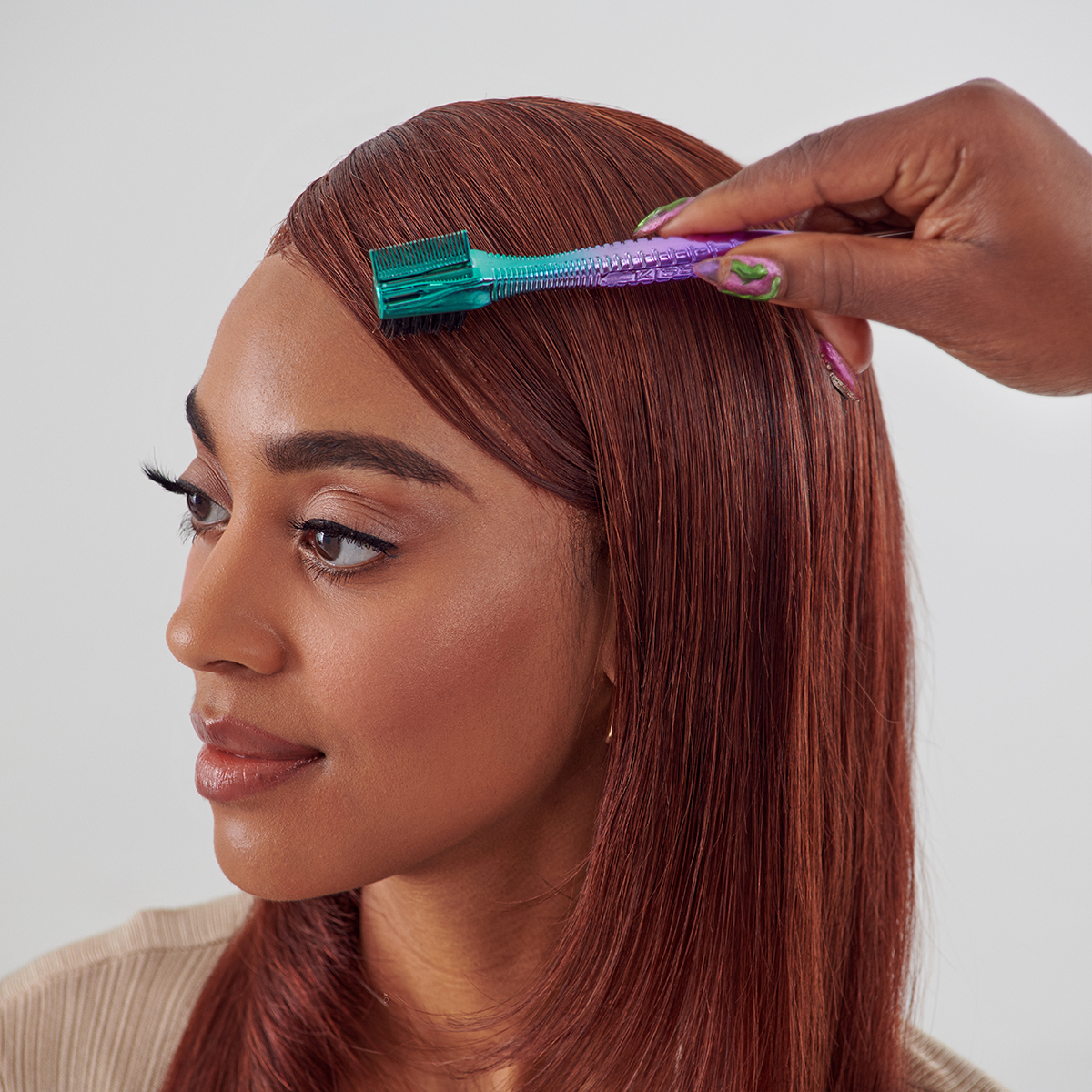Hand holding a small metallic ombre chrome 3-in-1 edge brush with green and purple colors, brushing the sleek side hair of a woman with straight reddish-brown hair.