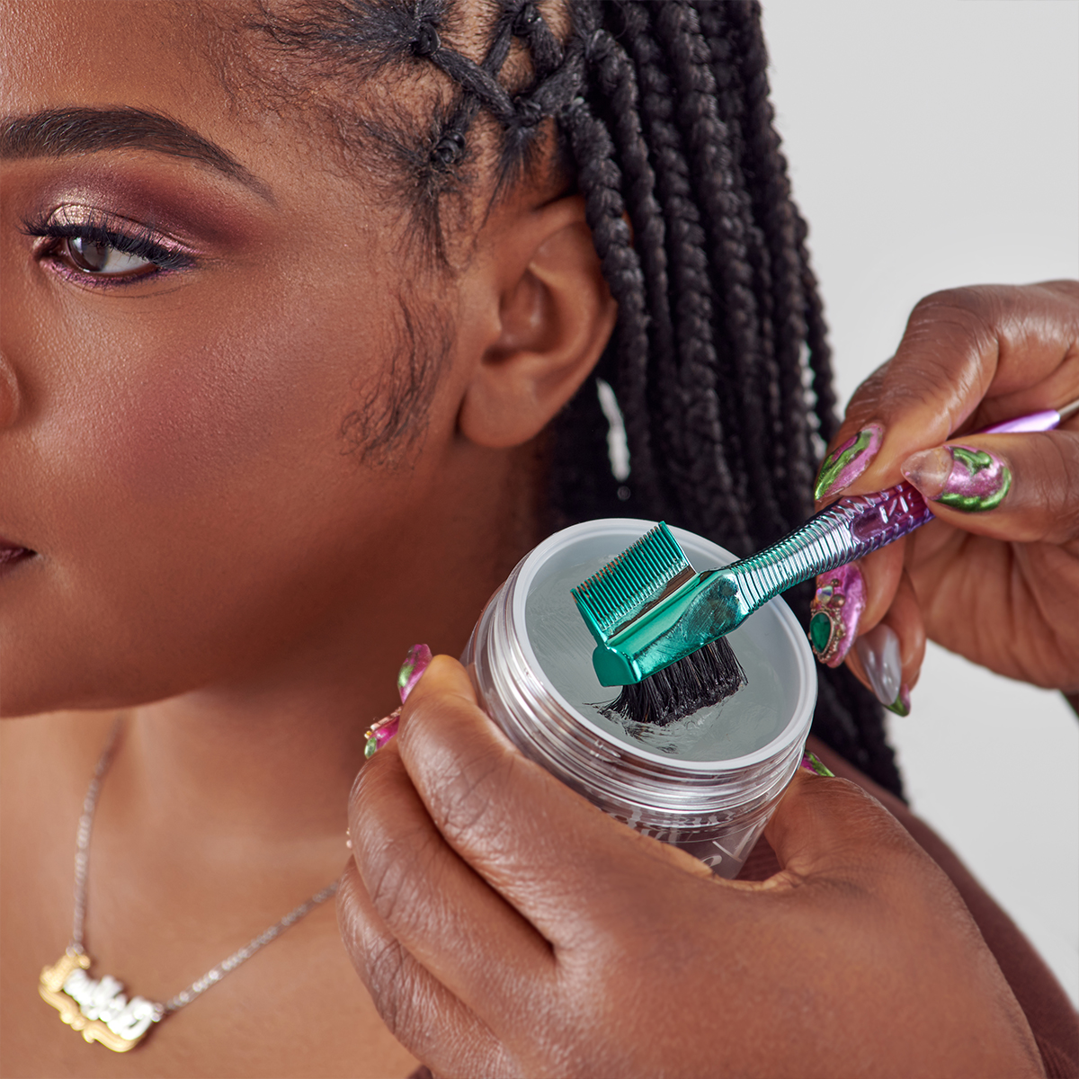 Close-up of a person with braided hair holding a clear jar of gel with turquoise comb-brush applicator dipped inside, preparing to style edges.