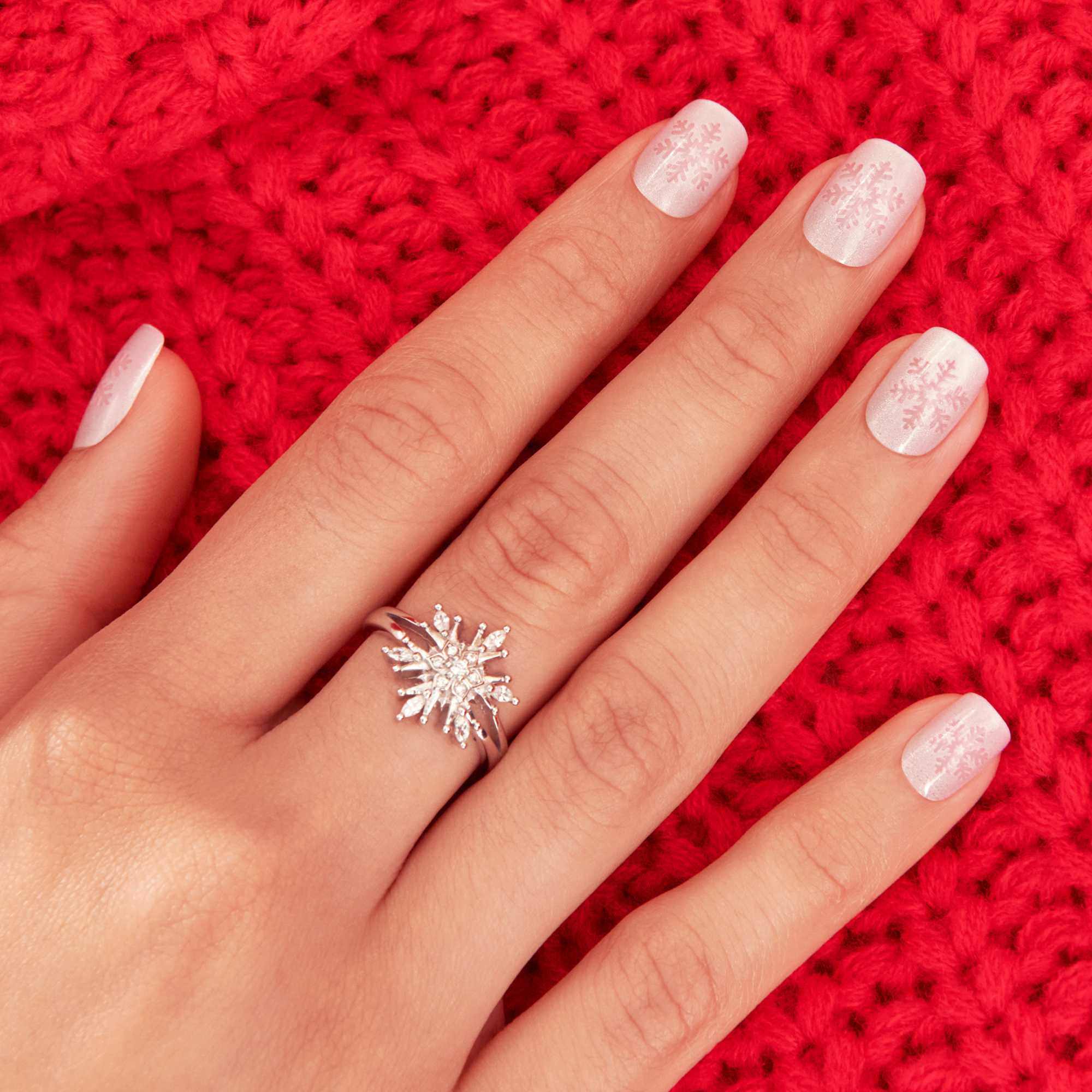 Hand with square-shaped, frosted white press-on nails featuring subtle pink snowflake designs, resting on a red knitted fabric background.