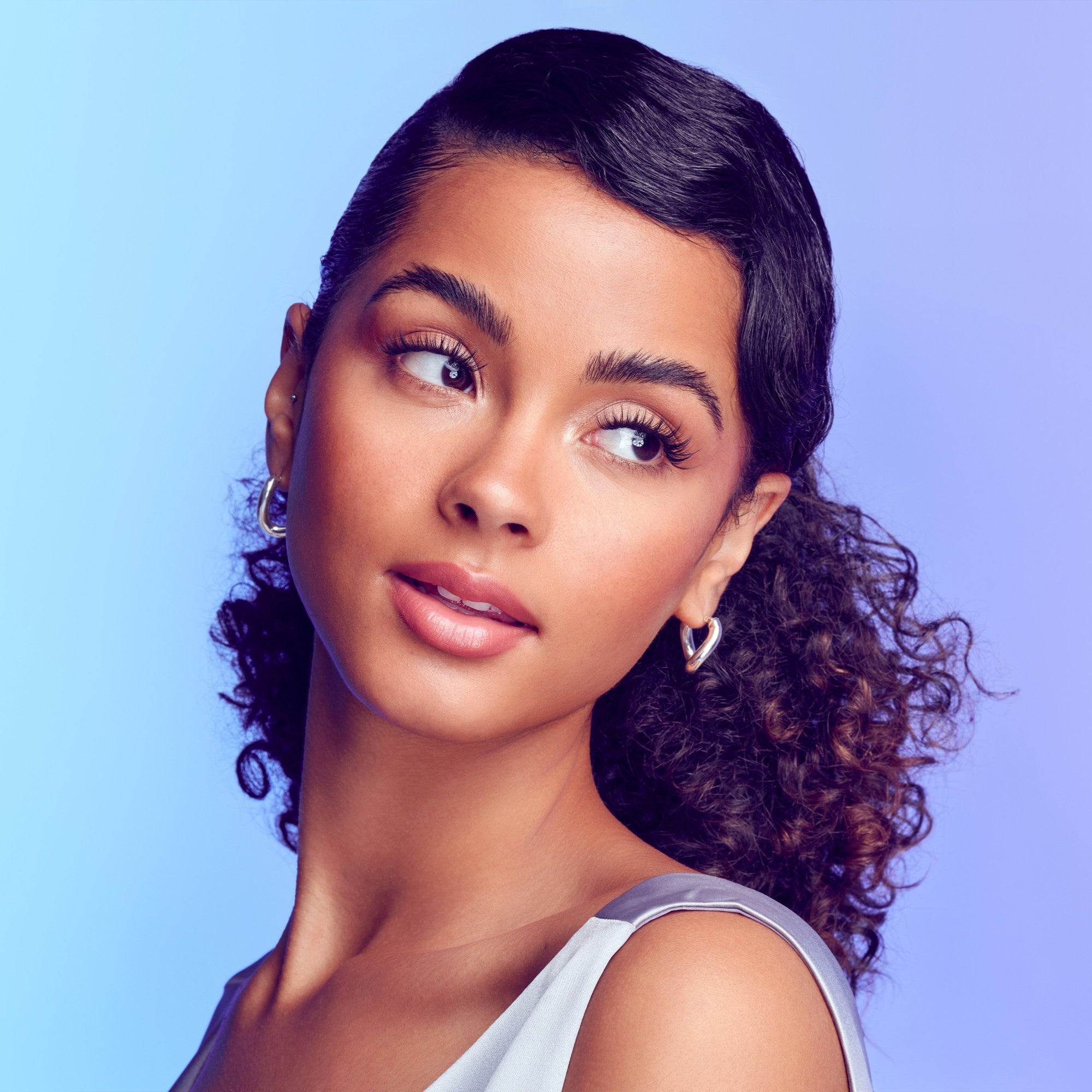 The image features a young woman with a graceful profile. She has a smooth complexion and her hair is styled with a combination of sleekness and soft curls. The background is a gradient of light blue and purple, creating a vibrant and modern atmosphere. Her expression is serene and thoughtful, accentuated by subtle makeup that highlights her features. She wears small hoop earrings, adding a touch of elegance to her look.