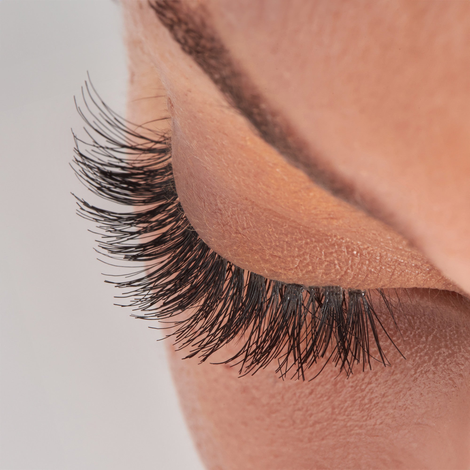 The image features a close-up view of an eye with long, thick eyelashes. The eyelid is closed, showcasing the lush lashes that extend outward. The skin around the eye appears smooth and has a natural tone, emphasizing the contrast between the dark lashes and the lighter eyelid. The focus is on the intricate details of the eyelashes, highlighting their length and texture.