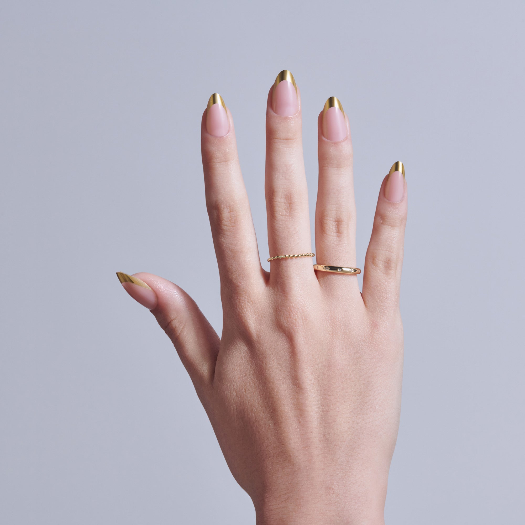 Hand displaying almond-shaped nails with pink base and gold tips, wearing two gold rings.