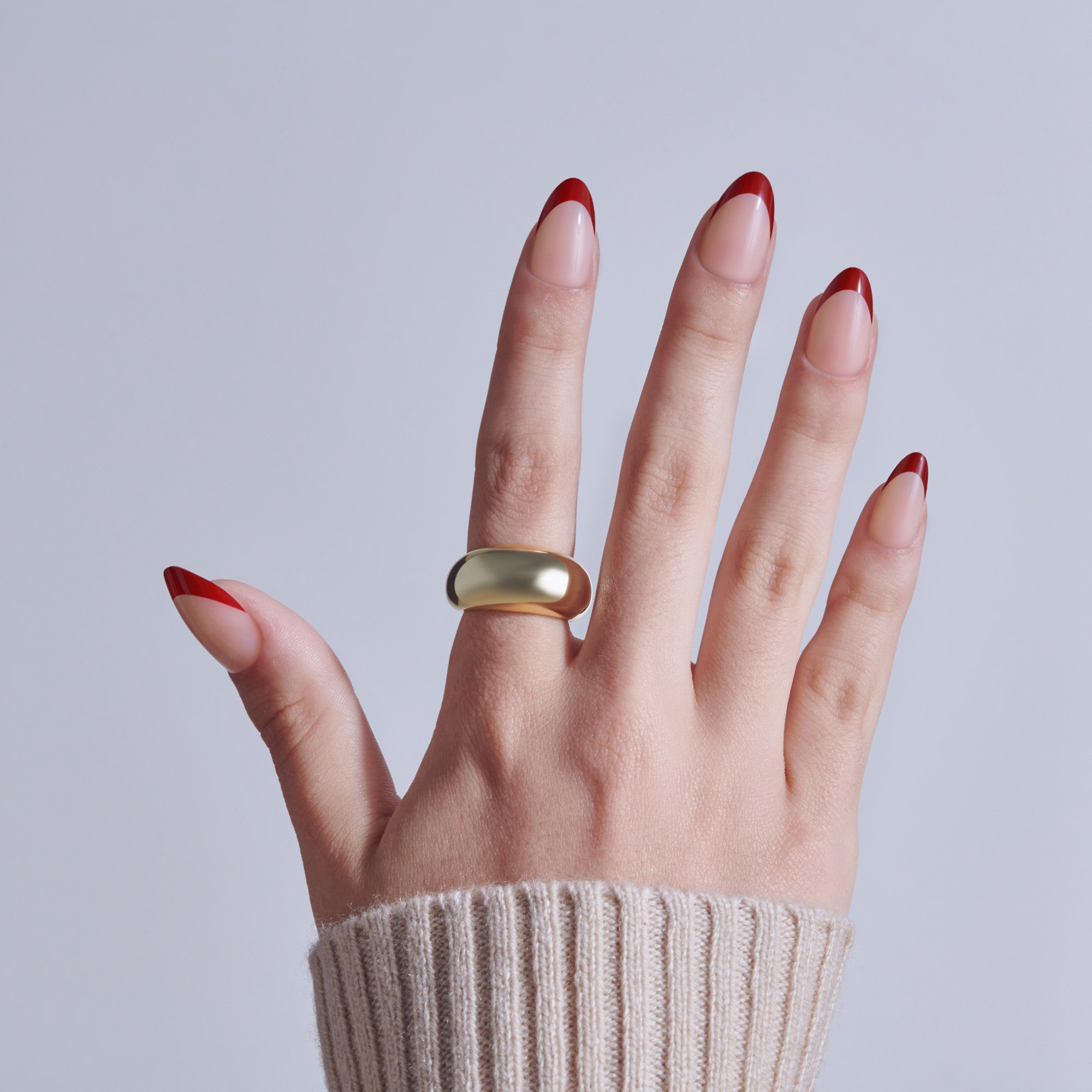 Hand with red French tip manicure and gold ring against gray background.