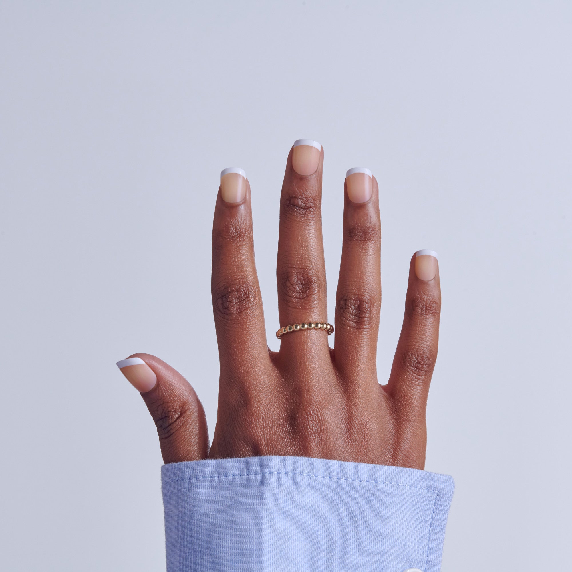 Hand with white French manicure nails and gold ring against blue sleeve on gray background.