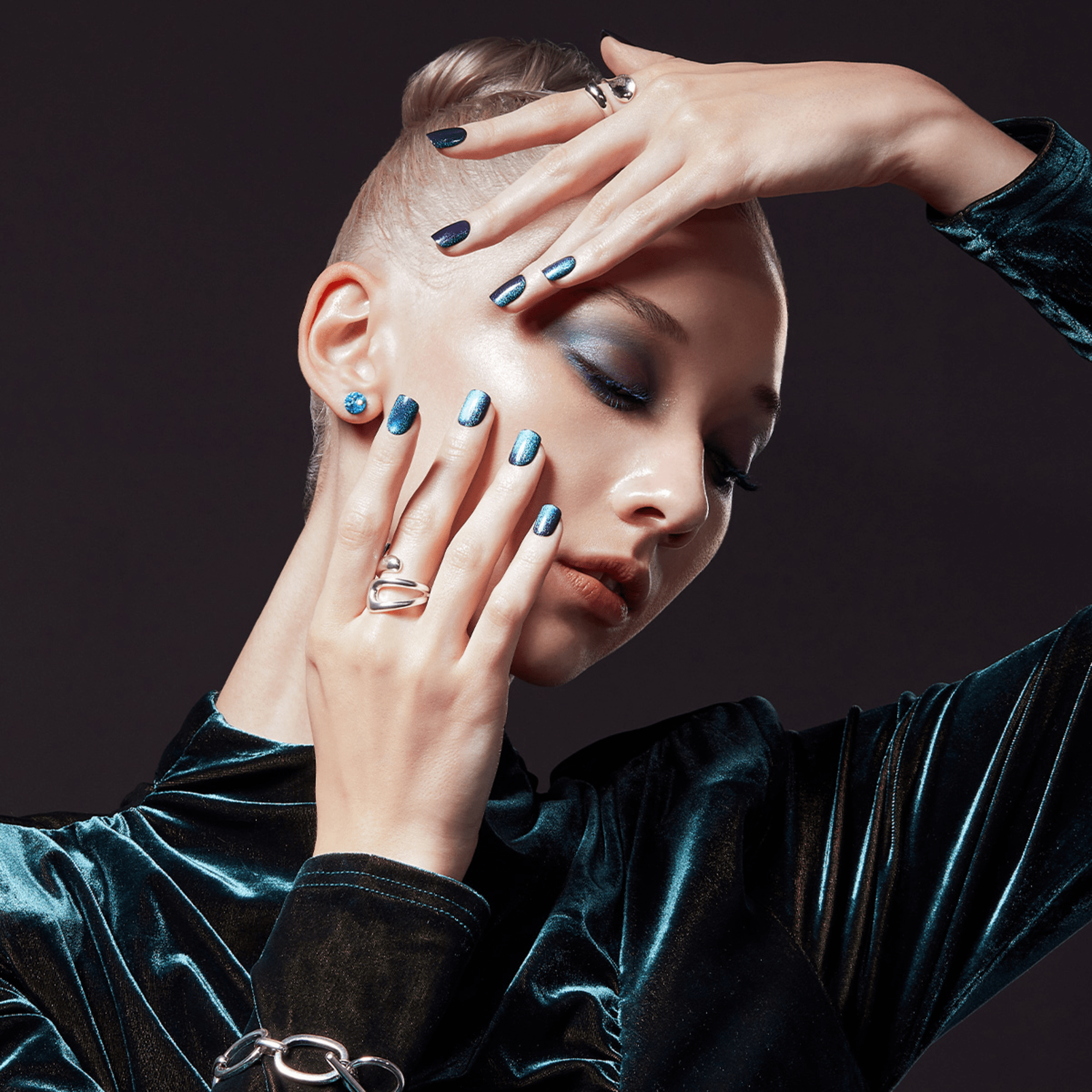 Person with shaved head wearing teal velvet, displaying blue metallic nail polish and silver rings.
