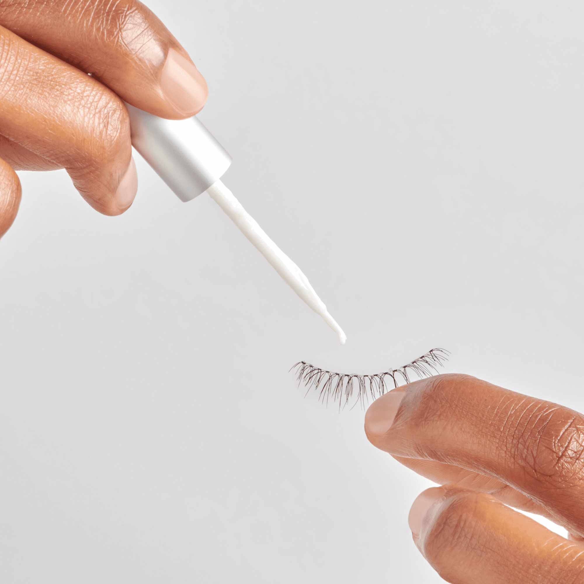 The image features a close-up of a hand holding a tube of adhesive, with a drop of white glue being dispensed from the nozzle. In the other hand, there is a pair of false eyelashes, showcasing their delicate, wispy design. The background is plain and light, emphasizing the focus on the hands and the beauty products.