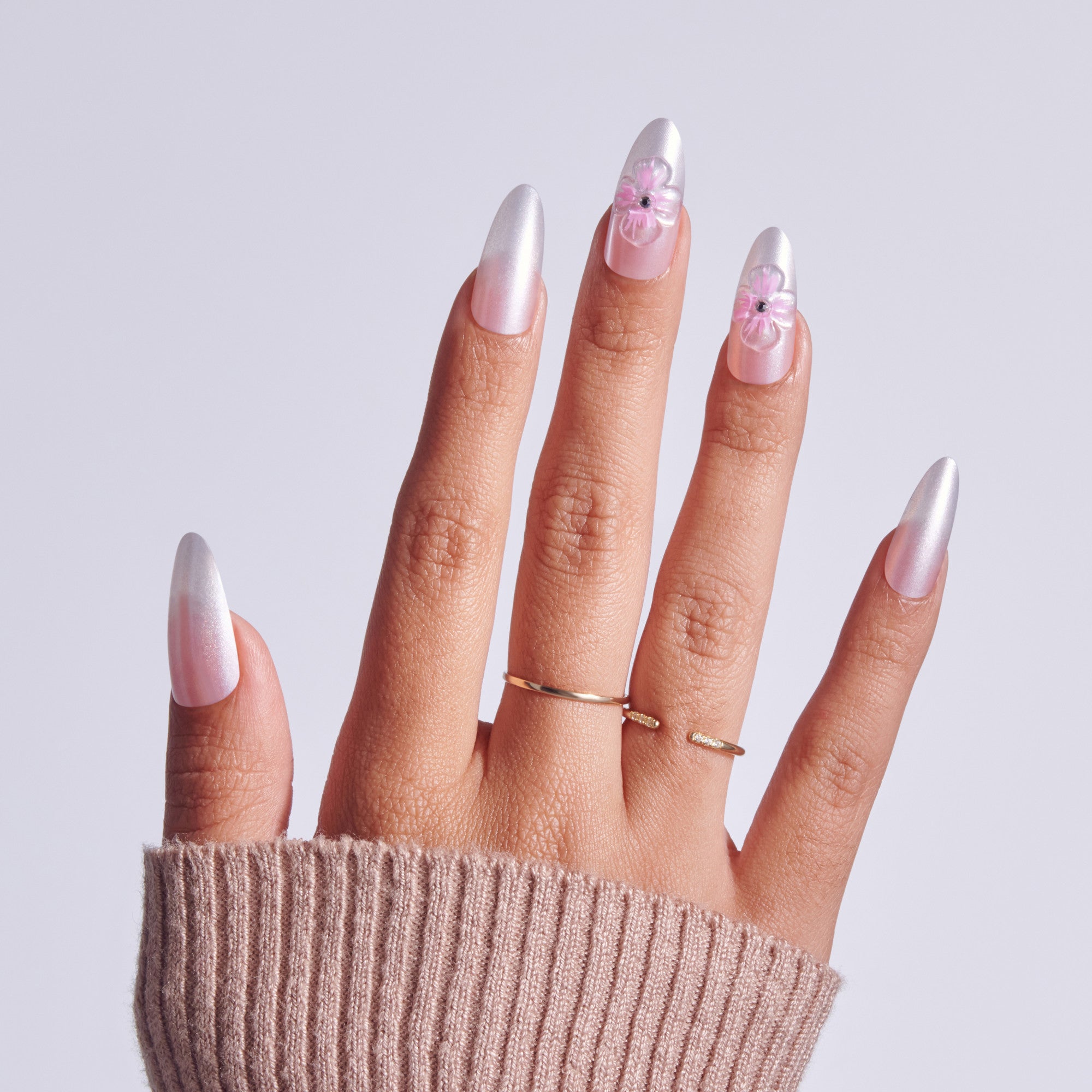 Long almond nails with pearl finish and pink floral designs, wearing gold rings.