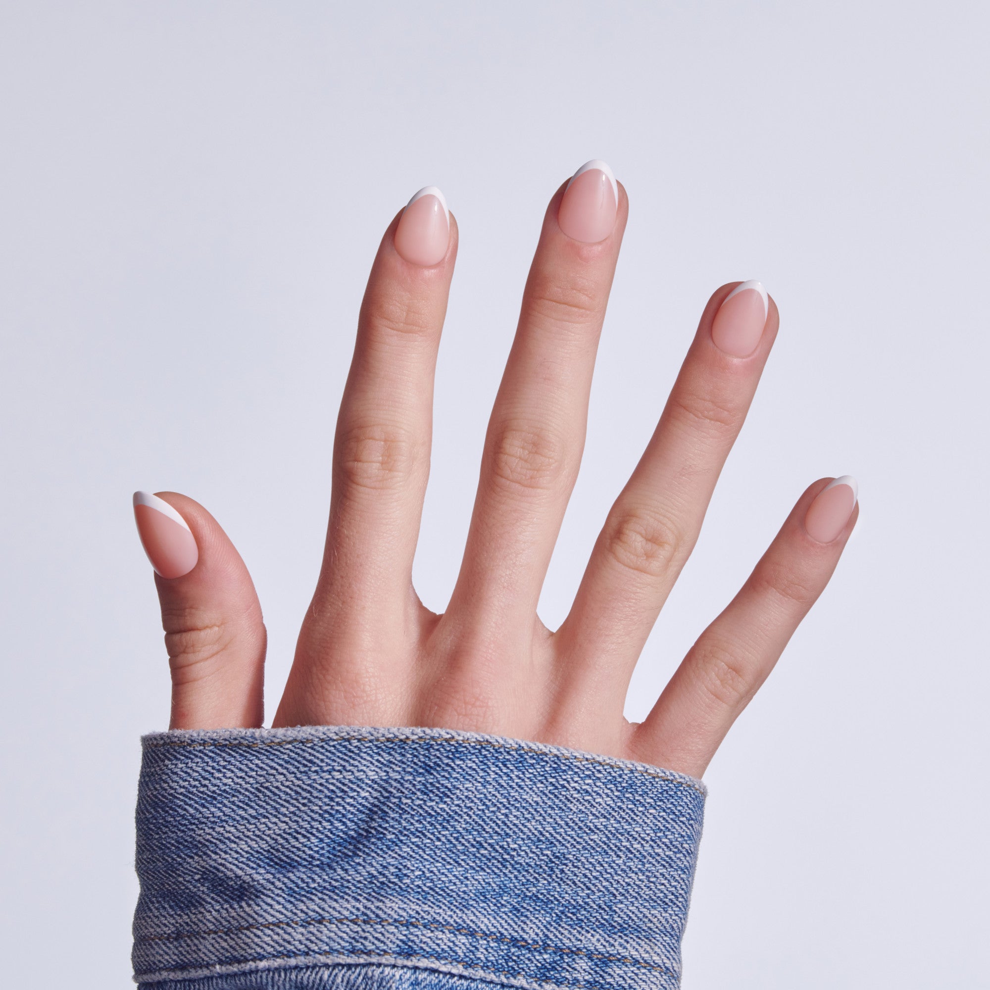 Hand with natural French manicure nails emerging from blue denim cuff against white background.