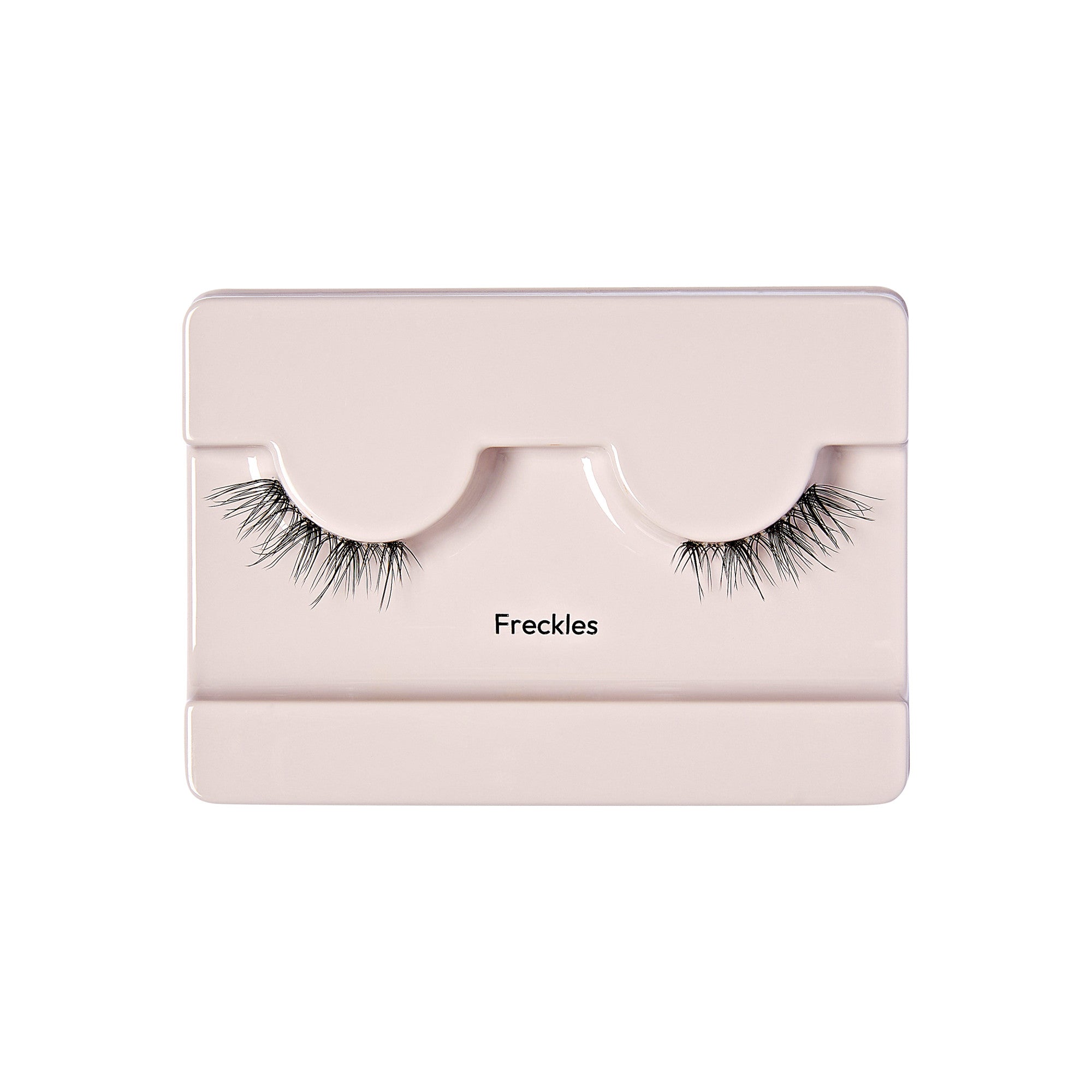 The image shows a pair of false eyelashes displayed in a light pink or beige plastic case. The case has two curved indentations where the eyelashes are placed, mimicking the shape of an eye. The eyelashes are long and wispy, with a natural look. Below the indentations, the word "Freckles" is printed in black text. The overall presentation is clean and minimalistic.