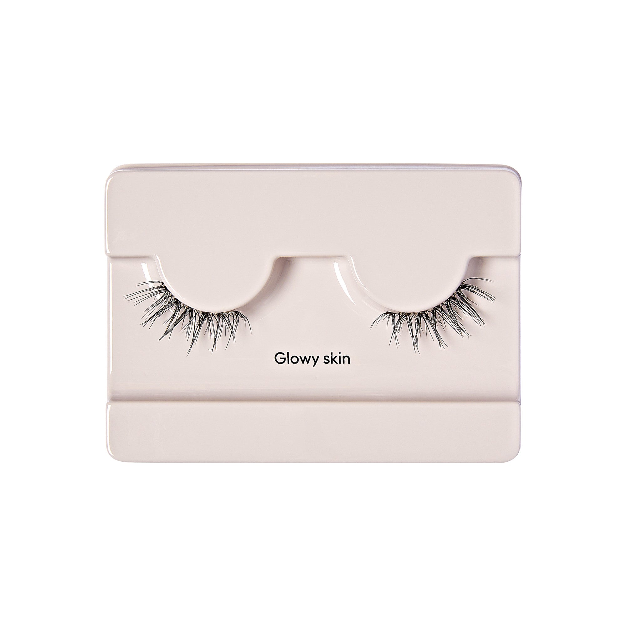 The image shows a pair of false eyelashes in a light-colored plastic packaging. The packaging is designed to hold the eyelashes securely, with two curved indentations where the lashes are placed. The false eyelashes are long and voluminous, with a natural-looking gradient from shorter to longer strands. The text "Glowy skin" is printed on the packaging, positioned between the two sets of eyelashes. The overall presentation is clean and minimalistic.