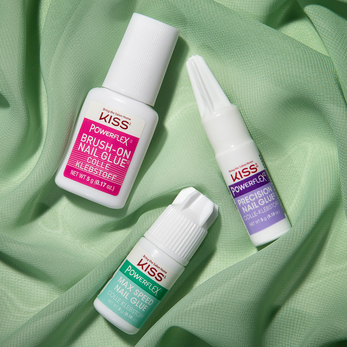 Three KISS PowerFlex nail glue bottles for different uses—Brush-On (pink label), Max Speed (green label), and Precision (purple label)—on light green fabric.