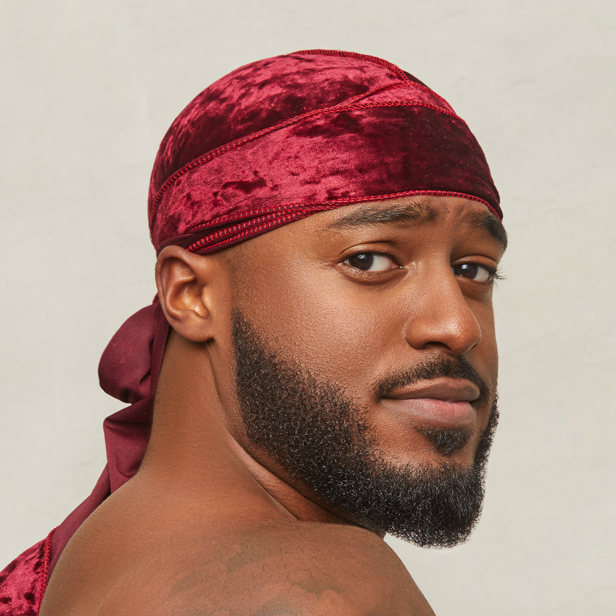 Close-up of a man wearing a burgundy velvet luxe durag, tied at the back, showing soft texture and rich color against a plain background.
