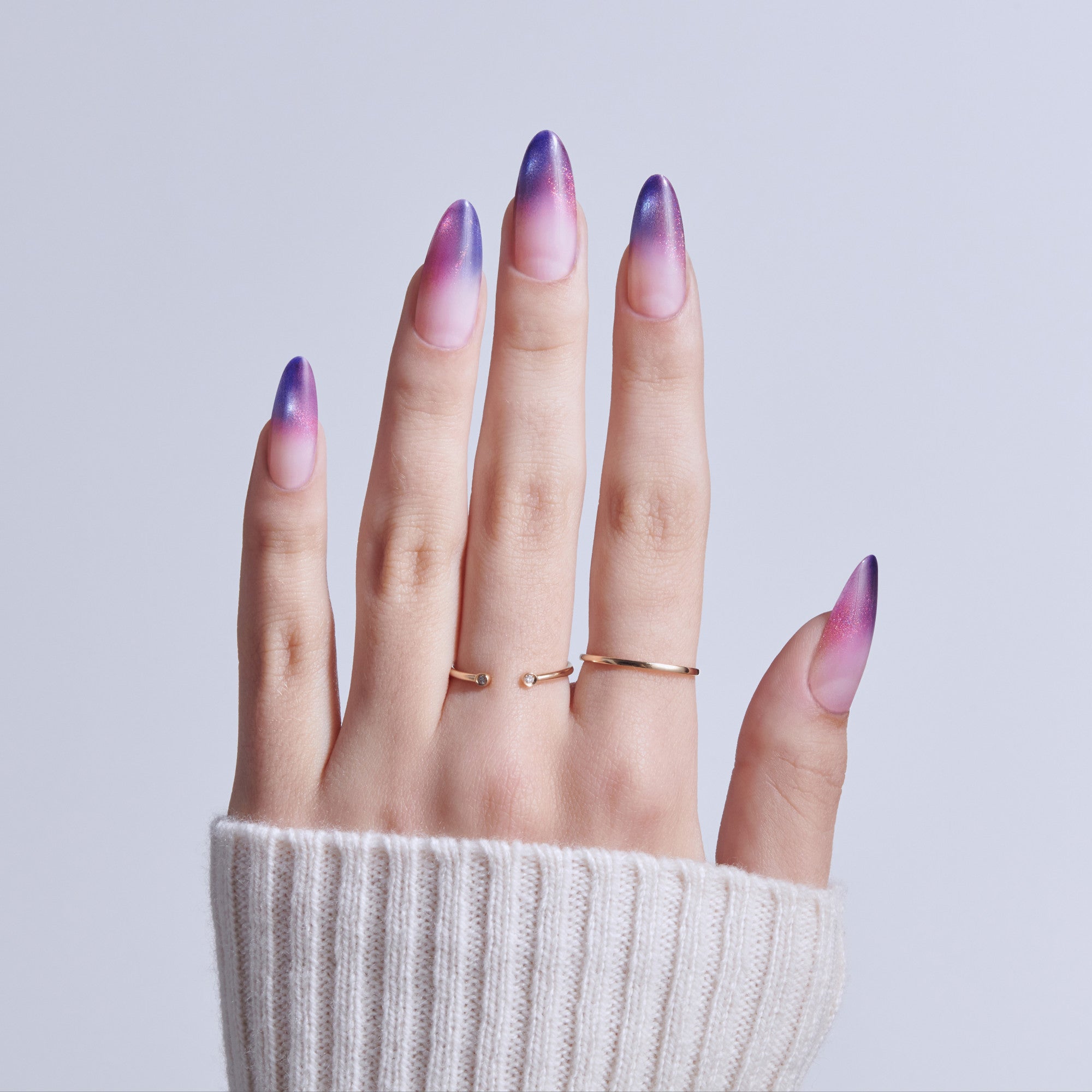 Hand with purple-to-pink ombre almond nails wearing gold rings against white background.