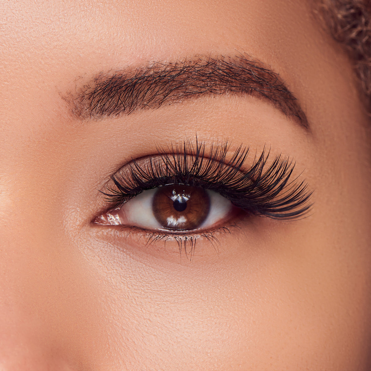 The image features a close-up of a human eye, showcasing a beautifully defined brow and long, voluminous eyelashes. The eye itself is a warm brown color, with a glossy surface that reflects light, enhancing its depth. The skin around the eye appears smooth and well-groomed, contributing to an overall polished look. The focus on the eye highlights its expressive nature and the meticulous attention to detail in the makeup, particularly the eyelashes.