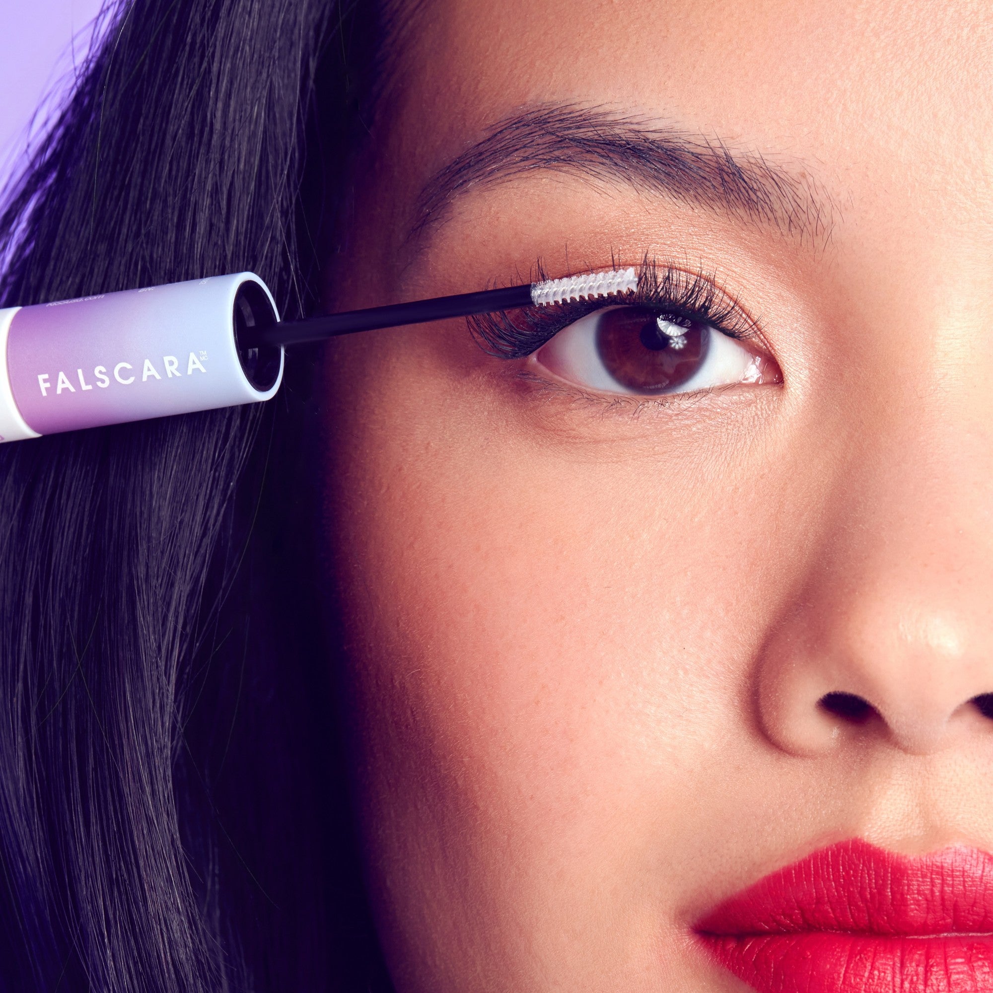 Close-up of a woman applying FALSCARA lash serum with a white brush wand to her upper eyelashes, showing long lashes and bright red lipstick.