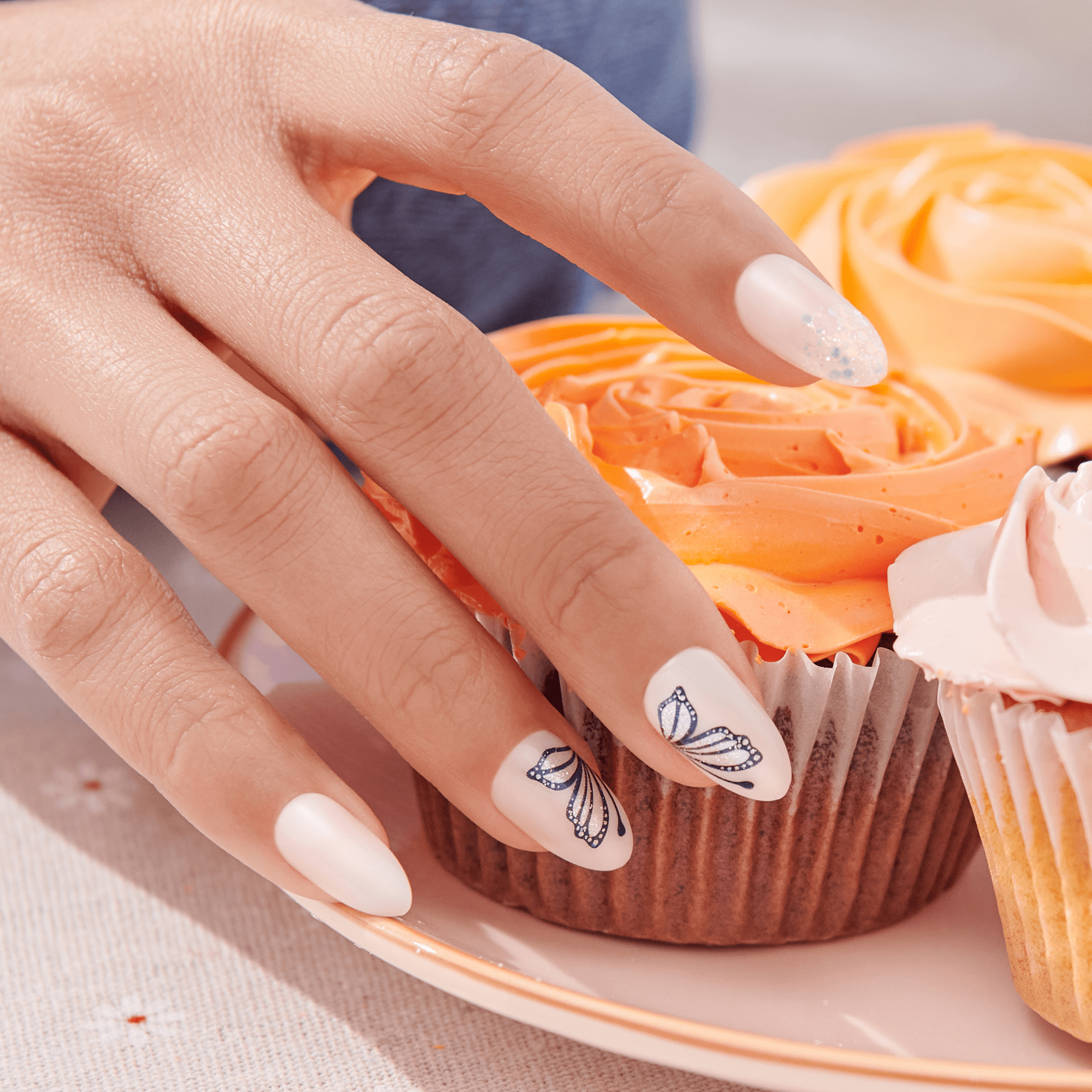 Hand with white press-on nails, two nails featuring delicate dark butterfly wing designs, one nail with glitter accents, resting on a plate with orange and pink frosted cupcakes.