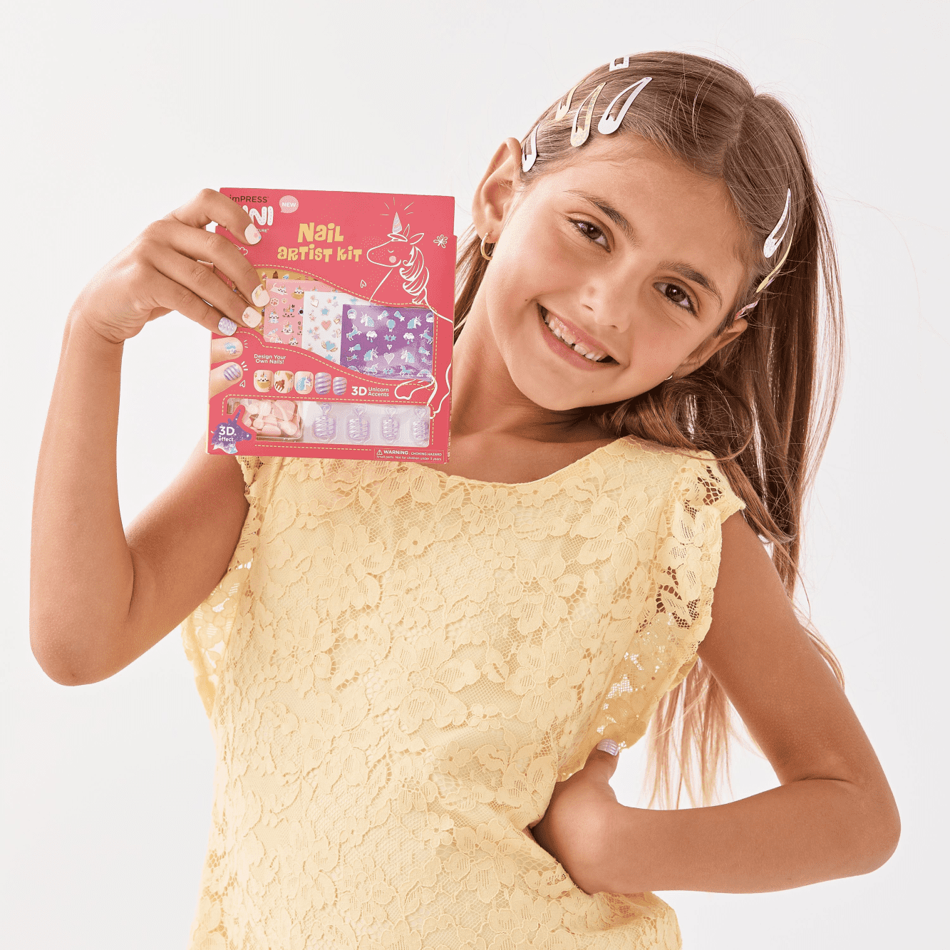 Smiling young girl in a yellow lace dress holds imPRESS MINI Nail Artist Kit packaging, featuring unicorn nail art with 3D accents and nail design samples.