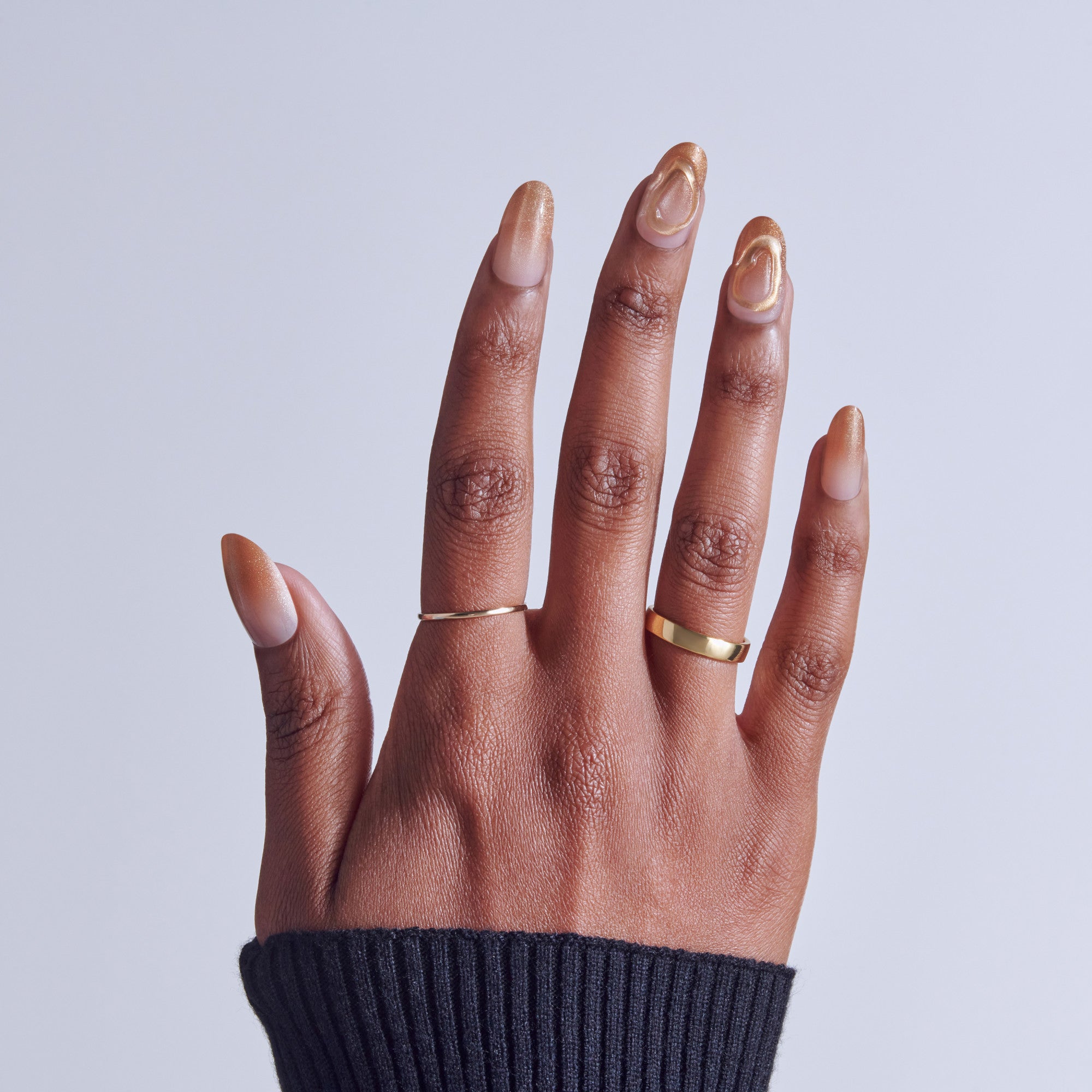 Hand with nude nail polish and gold rings against light background.