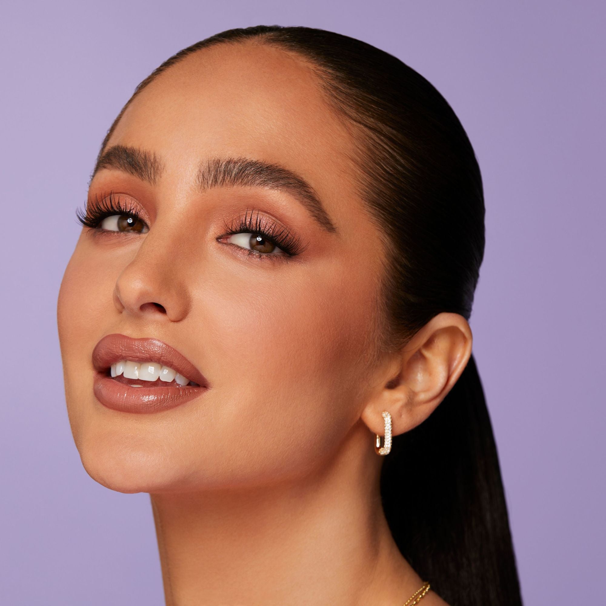 Close-up of a woman with smooth skin, wearing long, voluminous false eyelashes, neutral-toned eyeshadow, nude lipstick, and small gold hoop earrings against a lavender background.