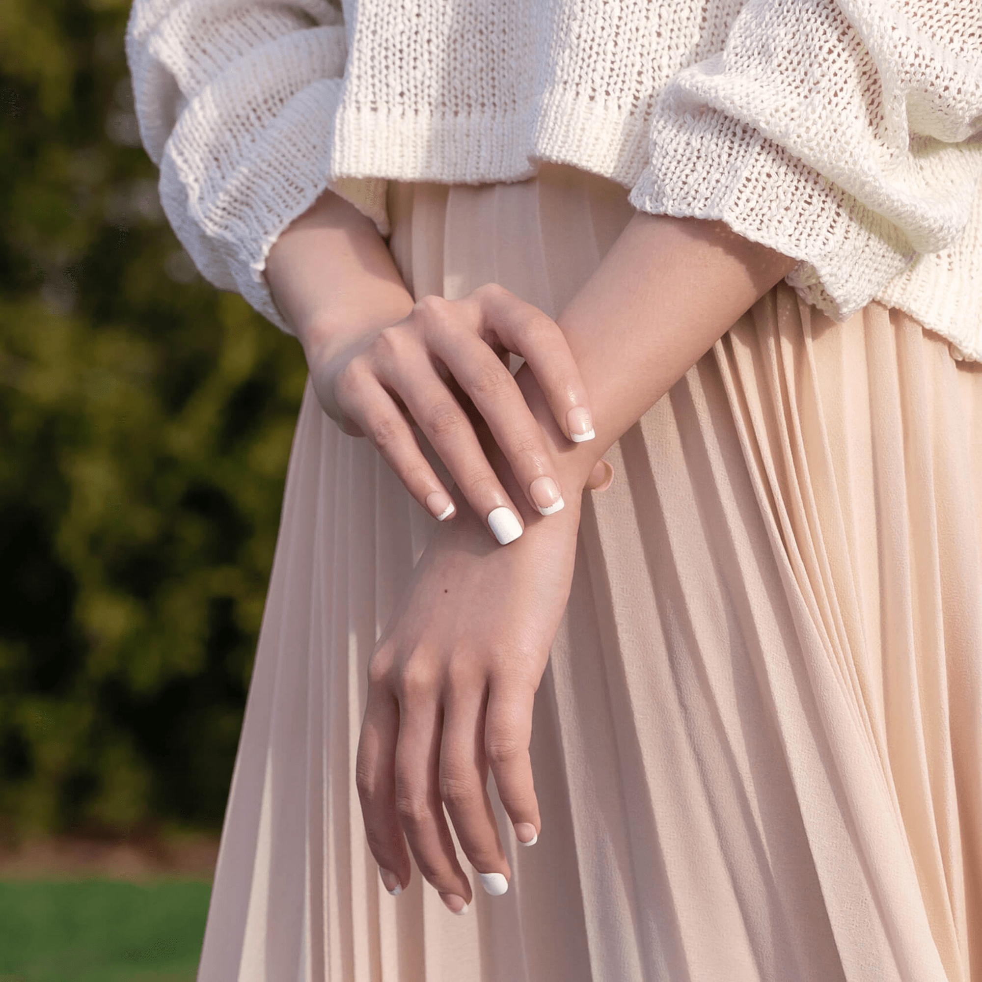 Hands with press-on nails featuring a mix of classic French tips and solid white designs, shown on a person wearing a cream sweater and pleated light pink skirt.