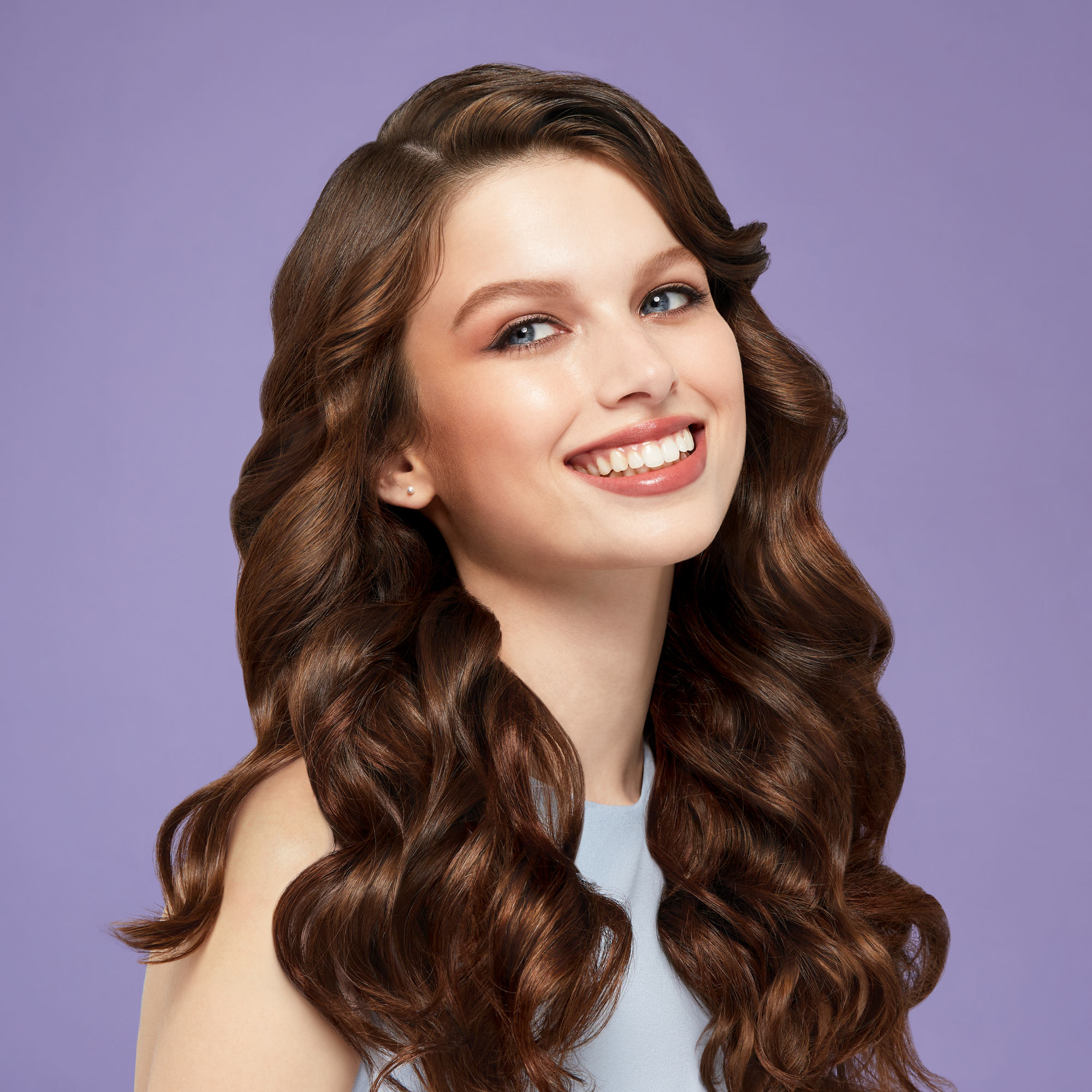 Smiling woman with long, shiny chestnut brown hair styled in smooth, voluminous 1-inch curls against a soft purple background.