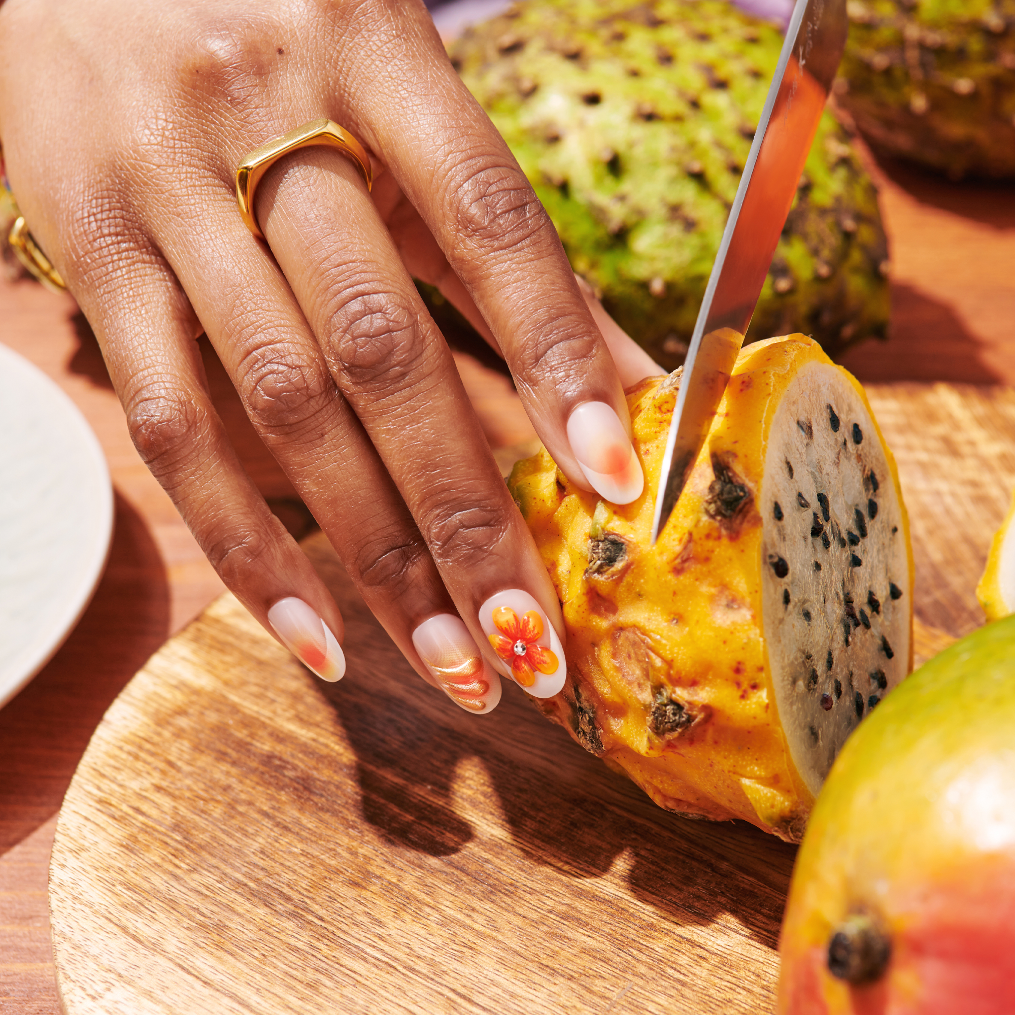 Hand with oval-shaped press-on nails featuring white base with orange floral nail art accents, gold ring on finger, holding sliced dragon fruit on a wooden cutting board
