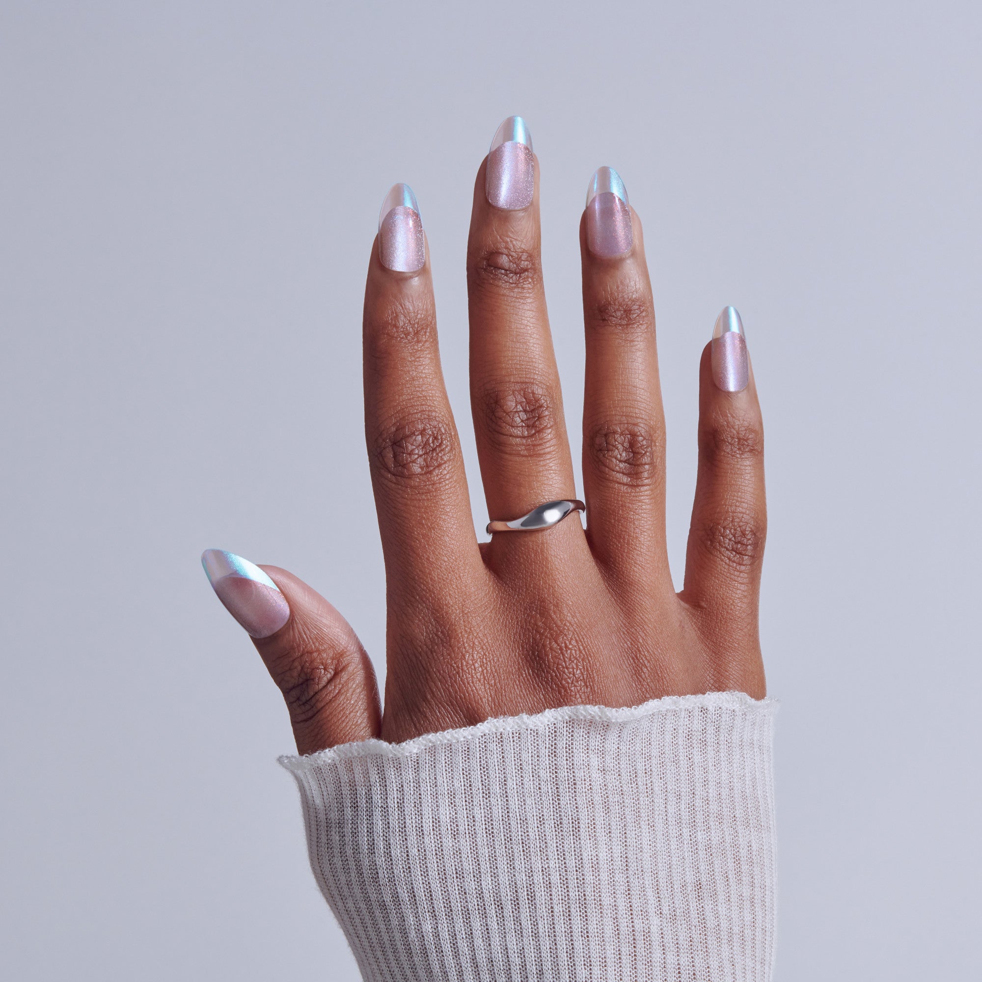 Hand with iridescent pink manicure and silver curved ring against white background.