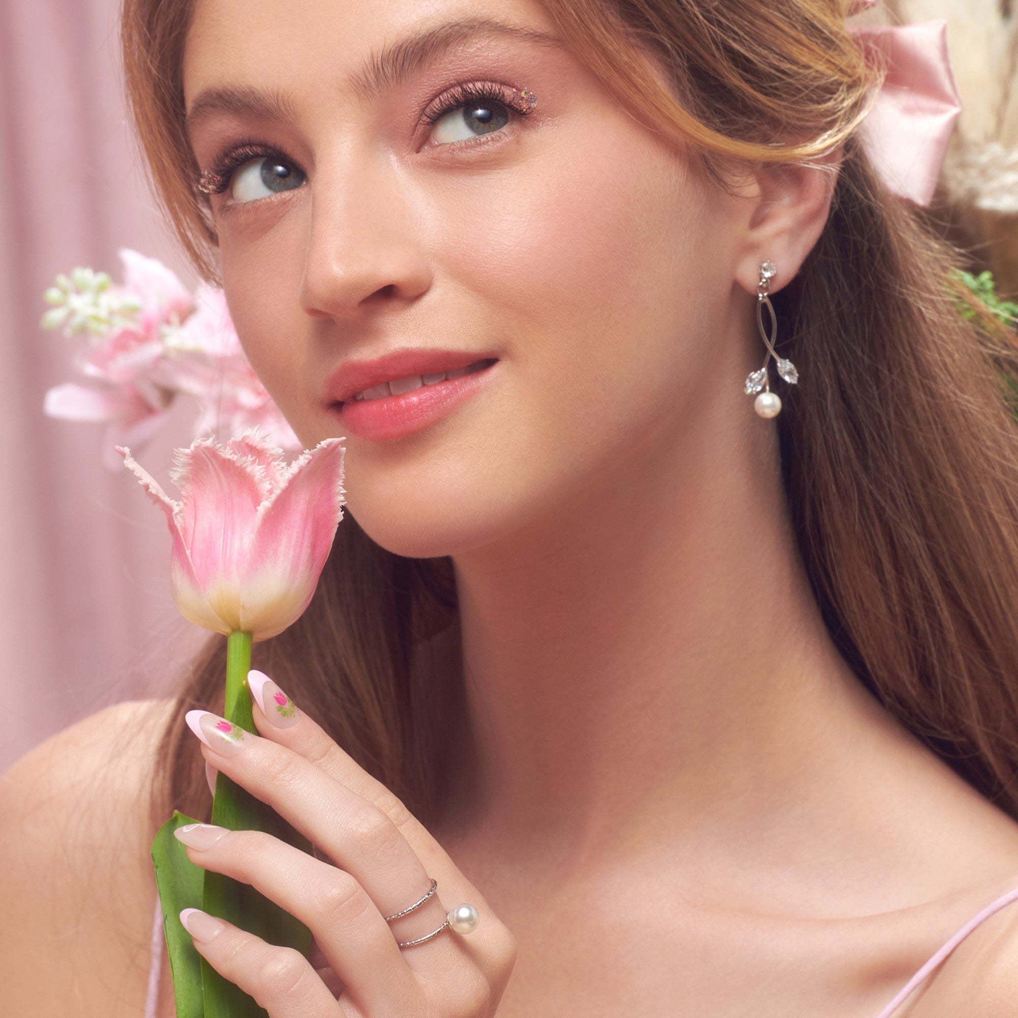 The image features a young woman with long, flowing hair, styled elegantly. She is holding a pink tulip close to her face, which adds a soft, romantic touch. Her makeup is delicate, with a subtle shimmer on her eyelids and a natural glow to her skin. She wears pearl earrings and a matching ring, enhancing her graceful appearance. The background is softly blurred with pastel colors, creating a dreamy atmosphere. The overall vibe is fresh and feminine, highlighting beauty and elegance.