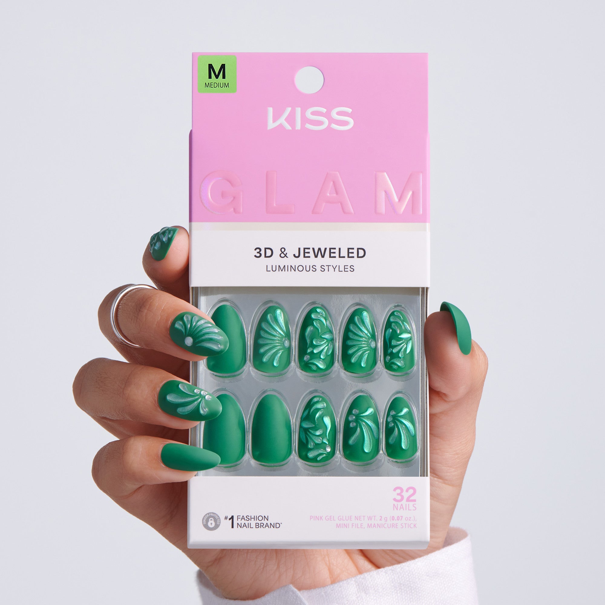 Hand holding KISS Glam 3D & Jeweled Luminous Styles press-on nails packaging, Medium size, 32 nails, dark green almond nails with white embossed floral designs on light gray background