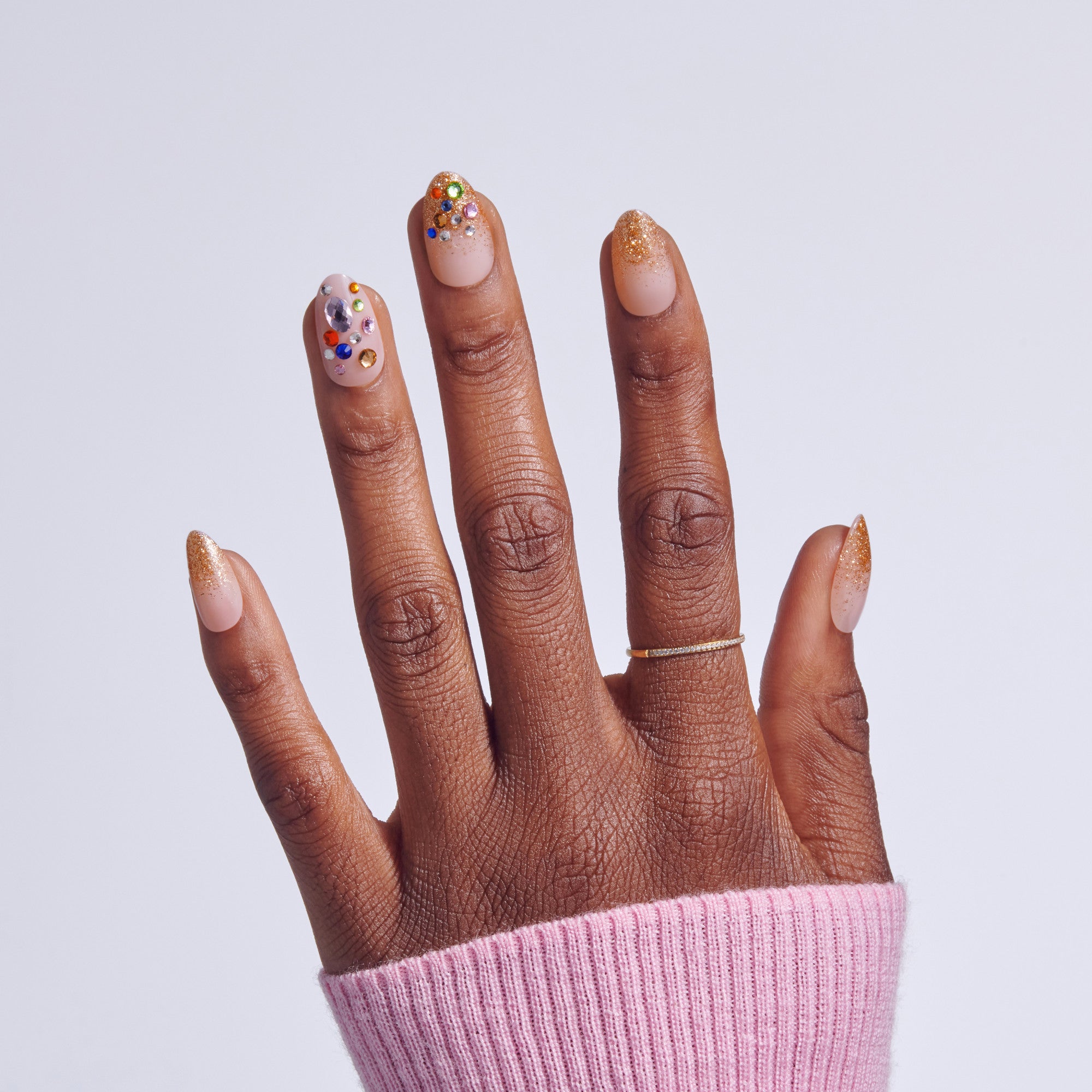 Hand with manicured nails featuring nude base, gold glitter tips, and colorful rhinestone accents.