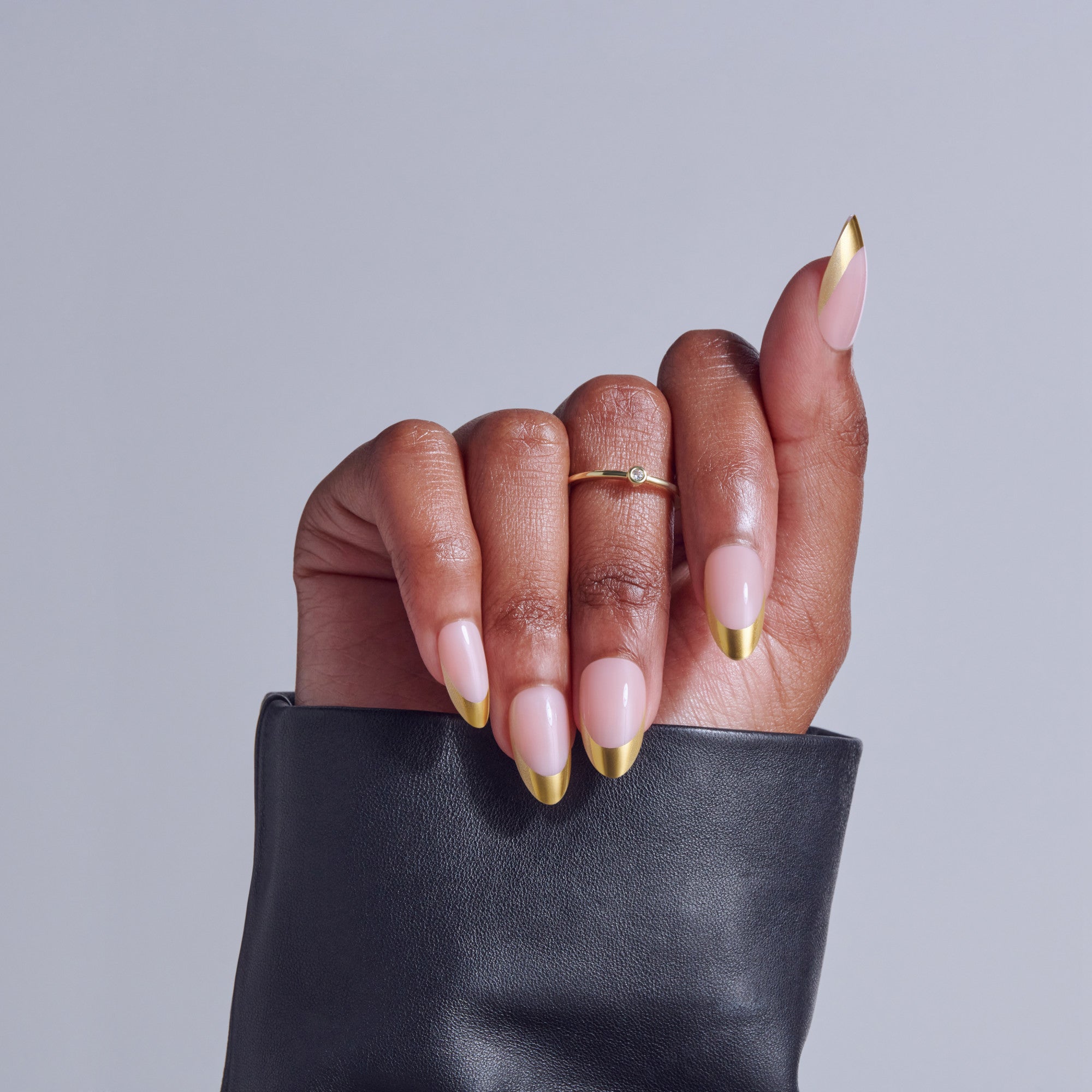 Hand with almond nails featuring pink base and gold tips, wearing gold ring, holding black leather clutch.