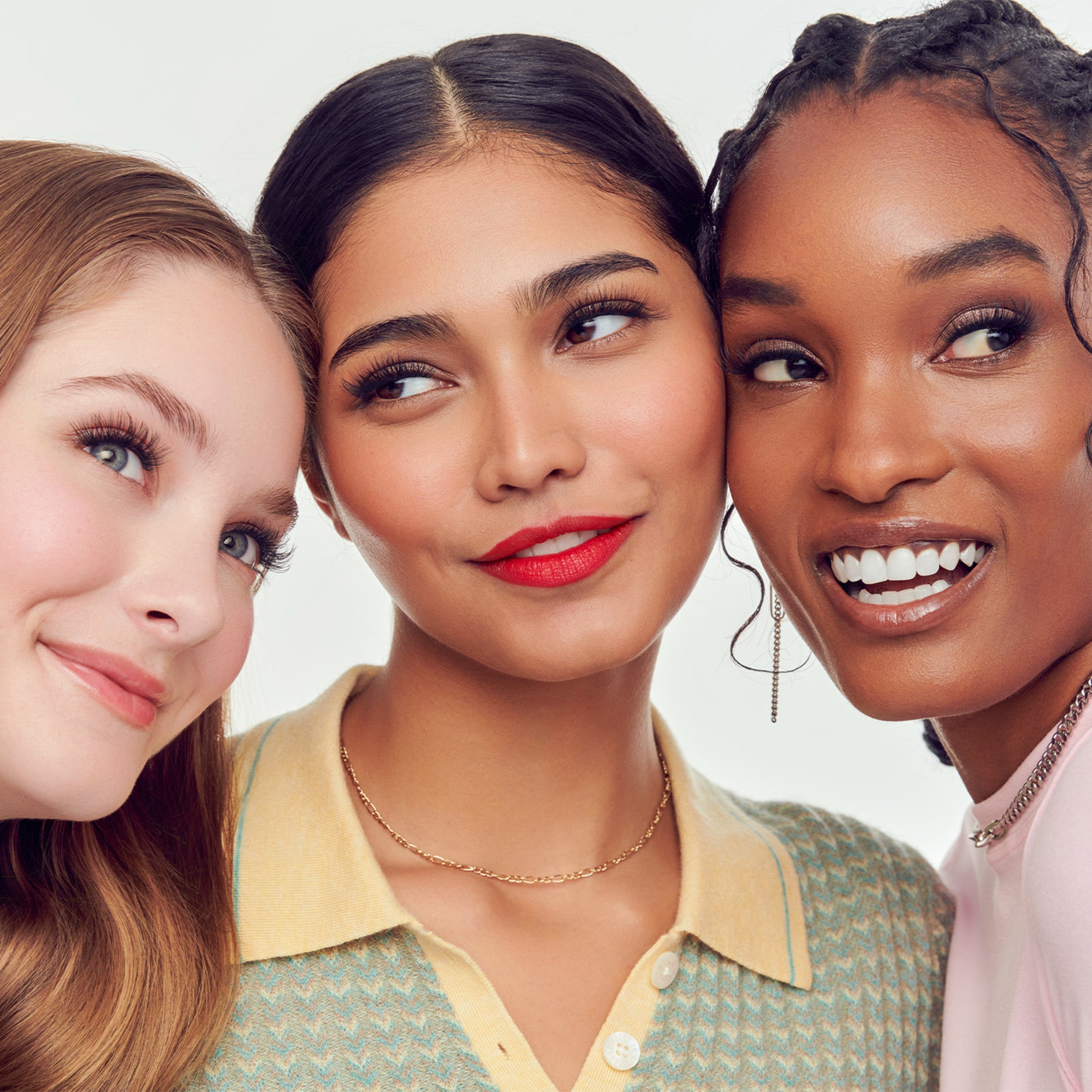 Three women with diverse skin tones smiling at camera, showcasing makeup and beauty looks.