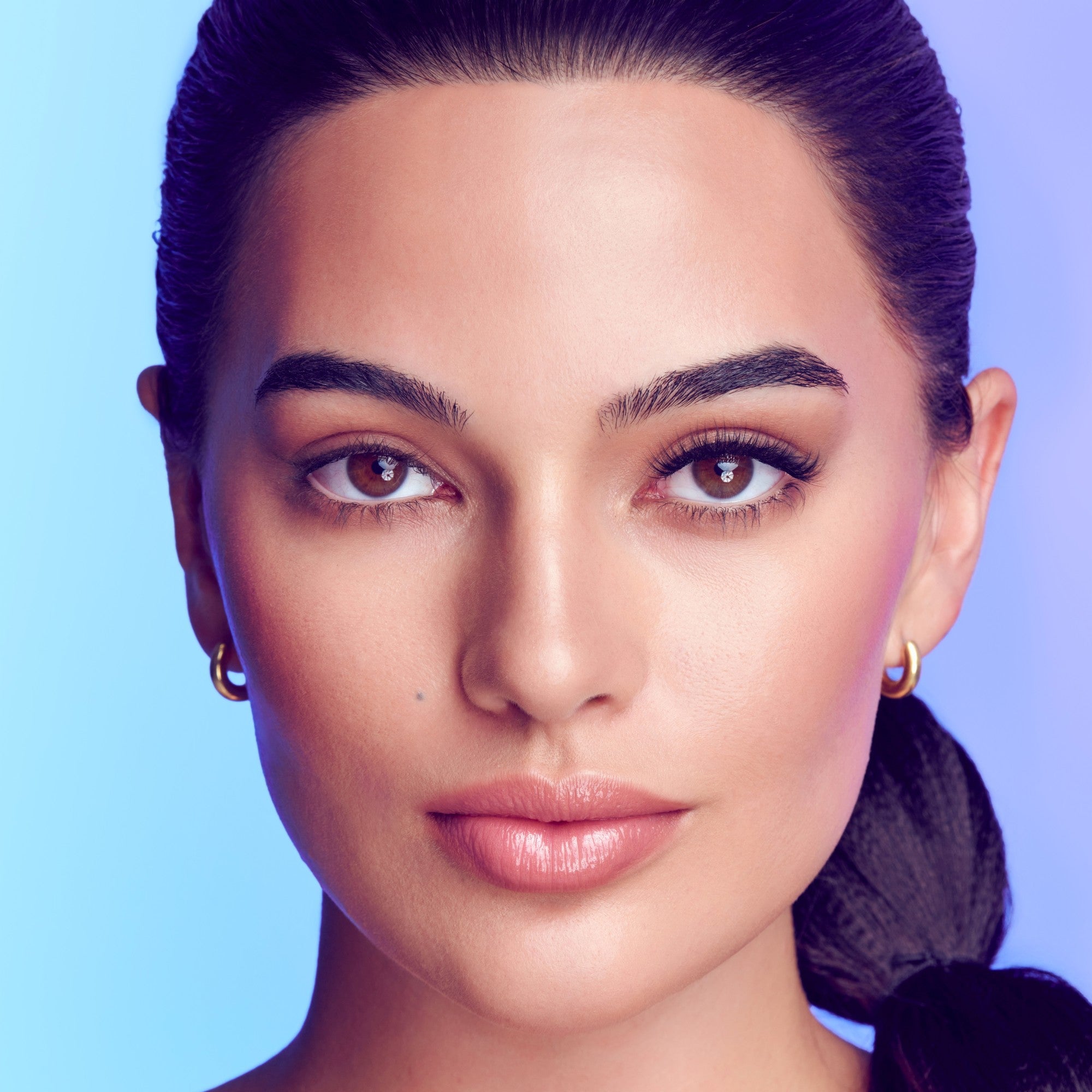 Close-up of a woman’s face against a blue-purple background, showing natural makeup with fuller, curled lashes on her right eye, highlighting eyelash enhancement.