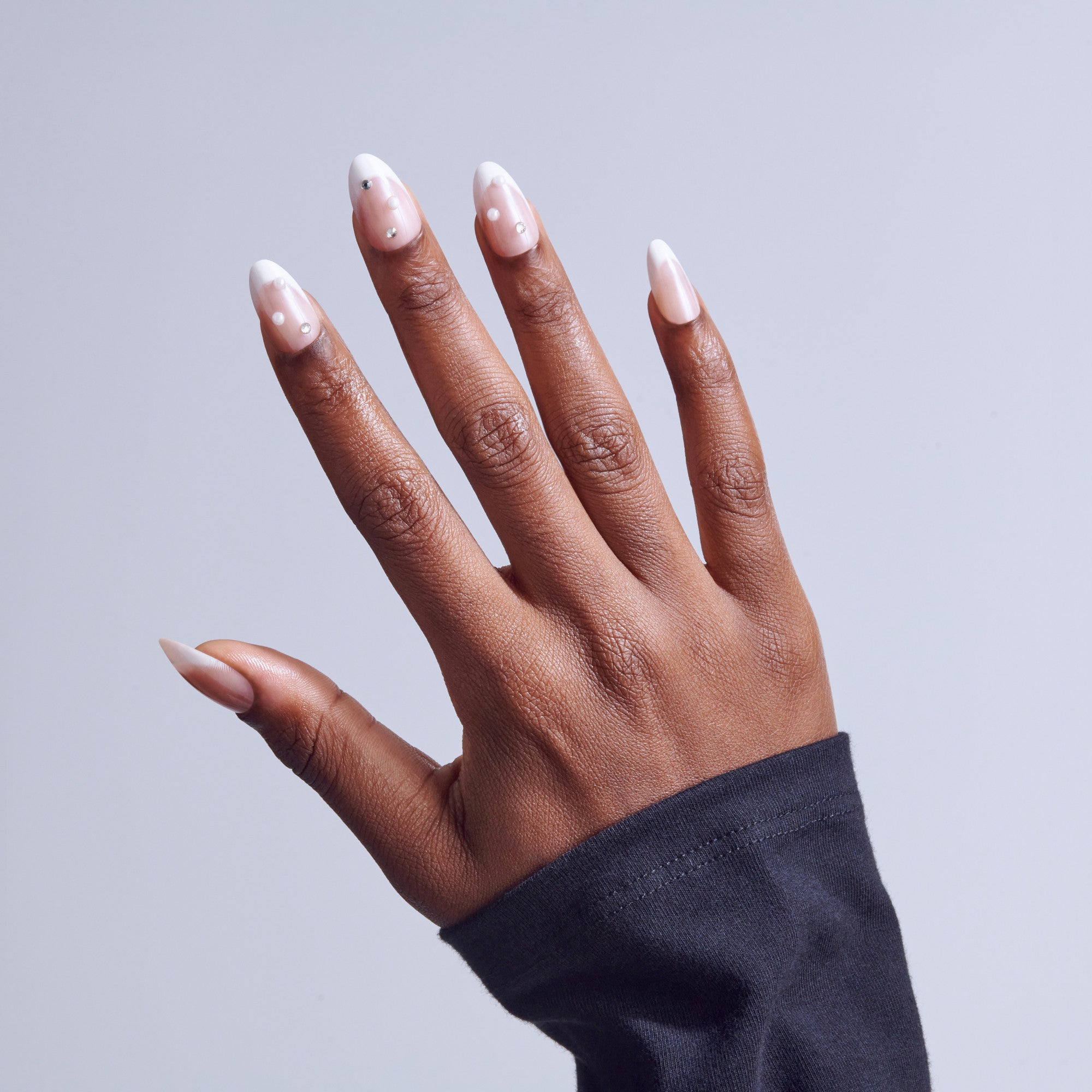 Hand with white-tipped almond nails and small white dots, wearing dark sleeve against gray background.