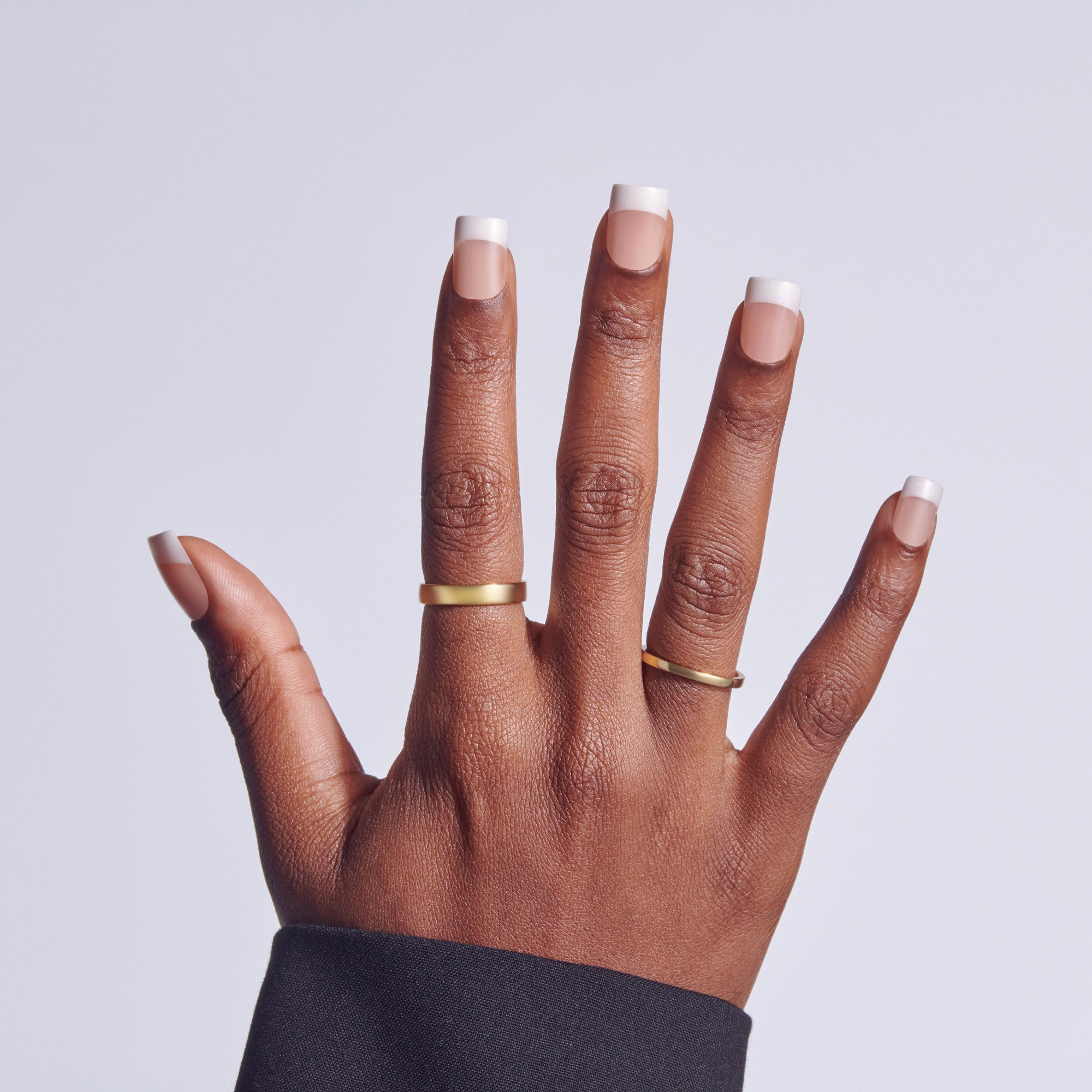 Hand with white-tipped manicure and gold rings against light background.