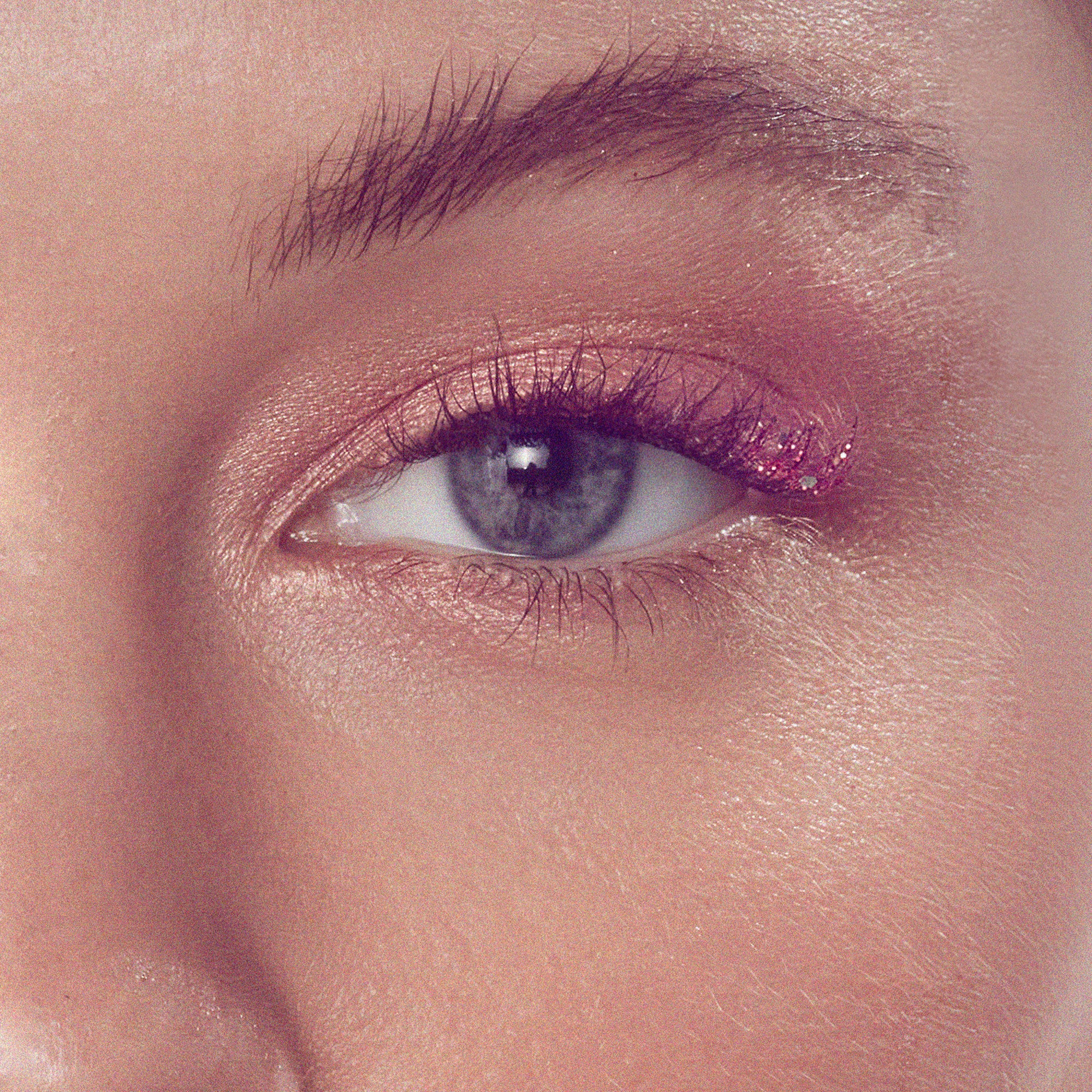 The image features a close-up of an eye with striking makeup. The eyelid is adorned with a soft, shimmery pink eyeshadow, while the lower lash line has a touch of glitter, adding a playful sparkle. The eye itself is a captivating shade of blue, framed by long, dark eyelashes. The skin around the eye appears smooth and radiant, contributing to an overall fresh and vibrant look.