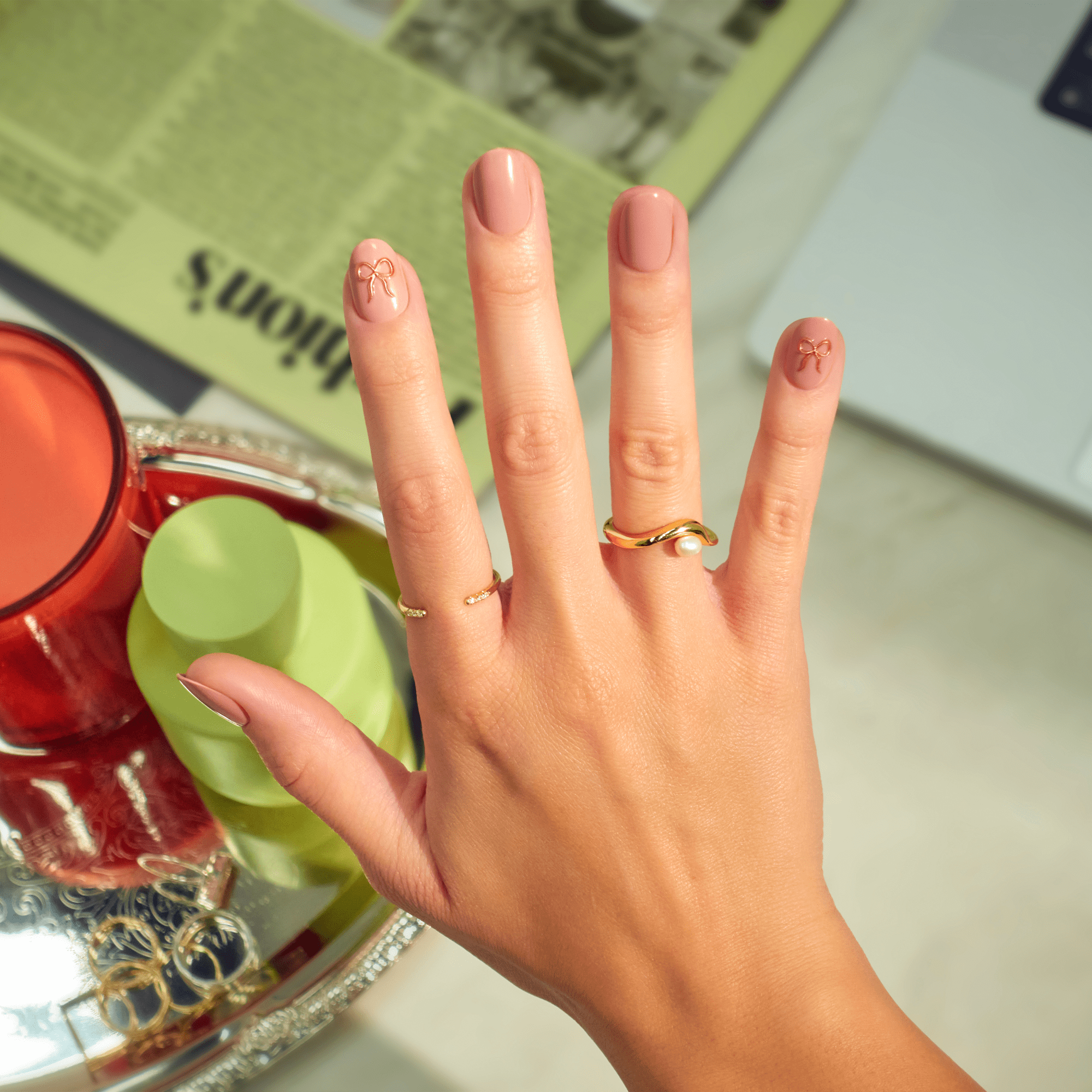 Manicured hand with pink nails featuring bow designs, gold rings, styled flatlay.