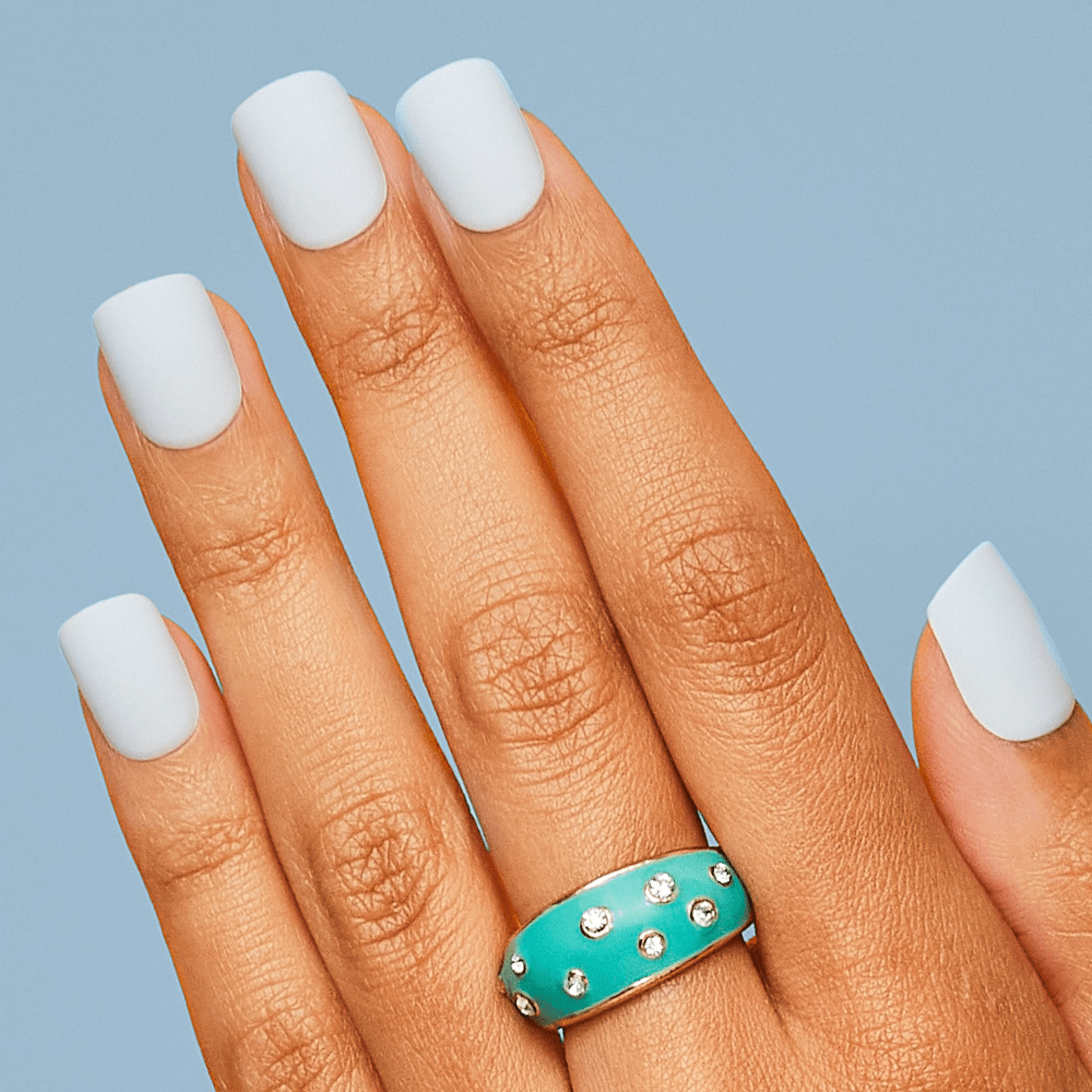 Hand with medium brown skin tone wearing white press-on nails with a matte finish against a light blue background. A turquoise ring with embedded crystals is on the ring finger.